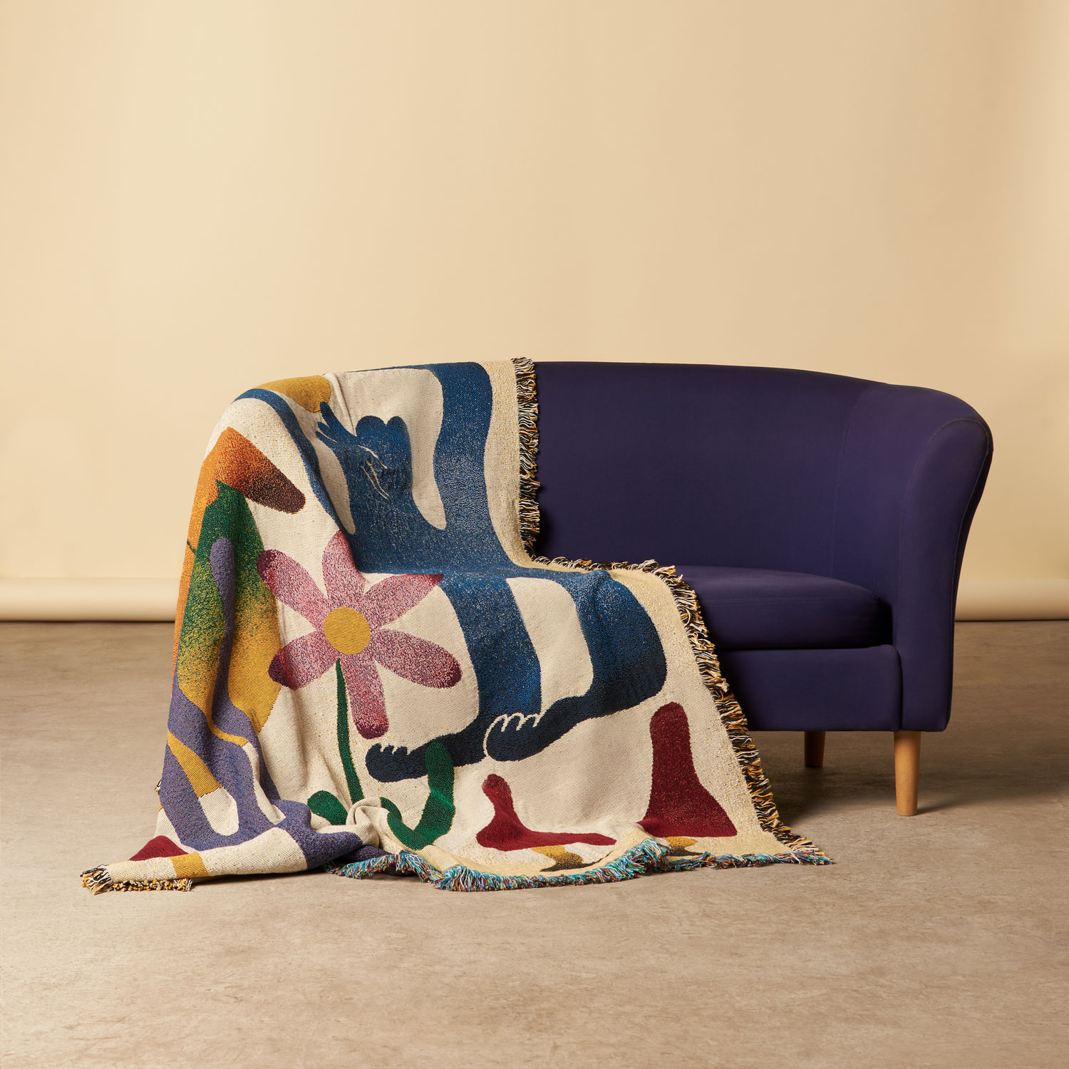 A purple sofa with a colorful blanket featuring floral and abstract patterns draped over it, against a plain background.