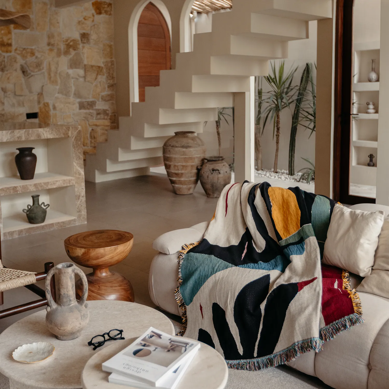 Living room with a sofa, blanket, and decorative items, staircase in the background, and a large window with plants.