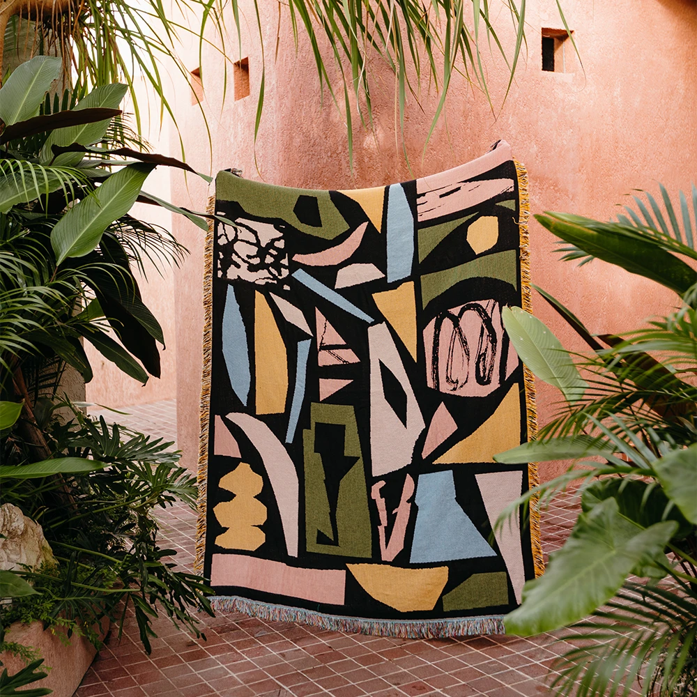 Colorful abstract patterned textile hanging among green plants against a pink wall.