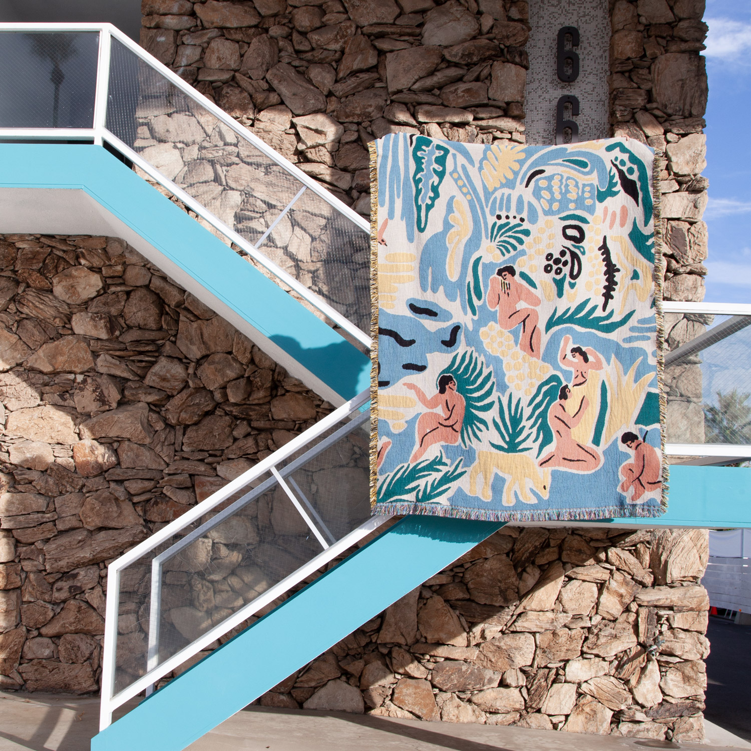 Colorful mural of people and plants on a large rectangular panel outside, with stone wall and blue staircase nearby.
