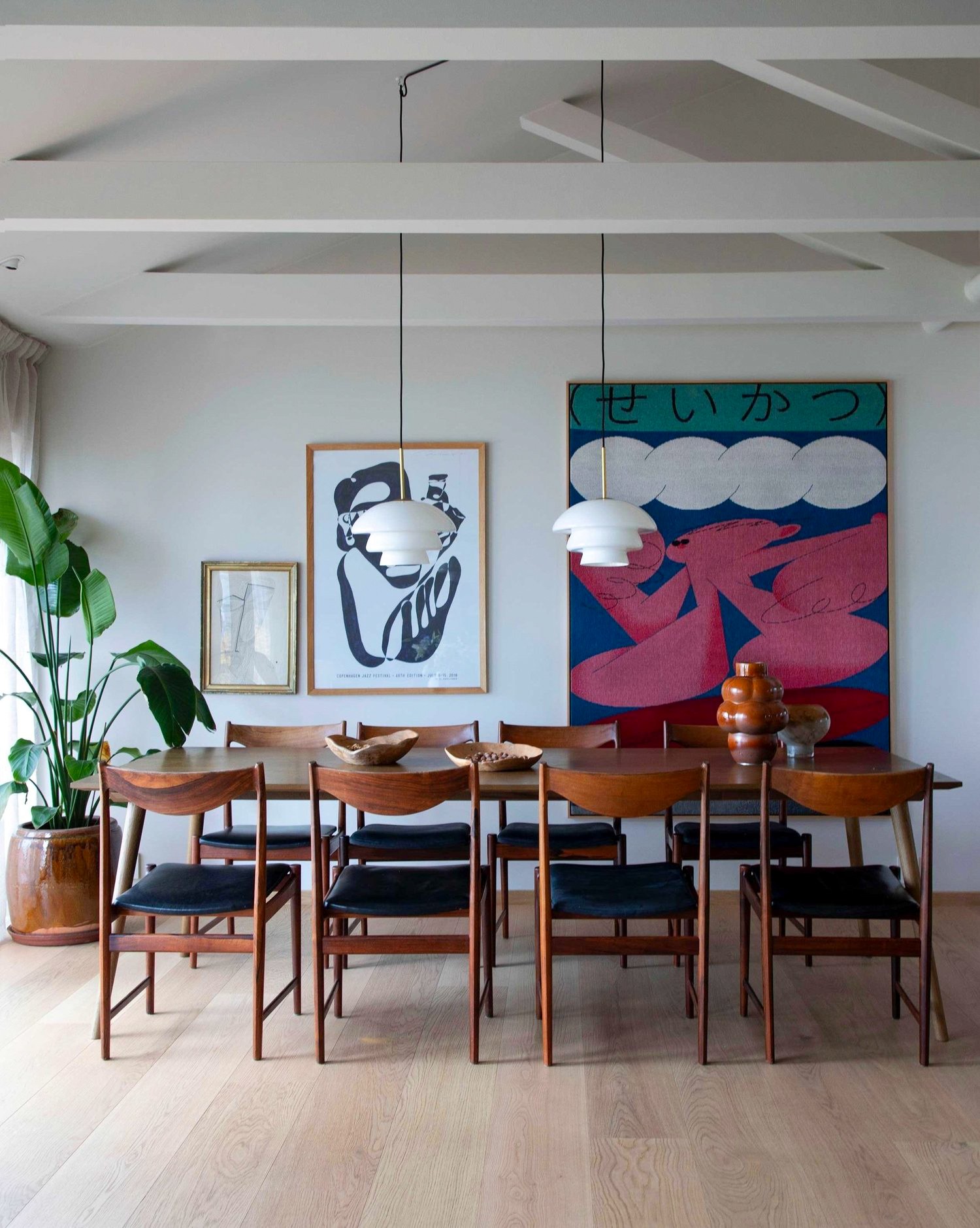 Dining room with wooden table, six chairs, large potted plant, artwork, and hanging pendant lights.