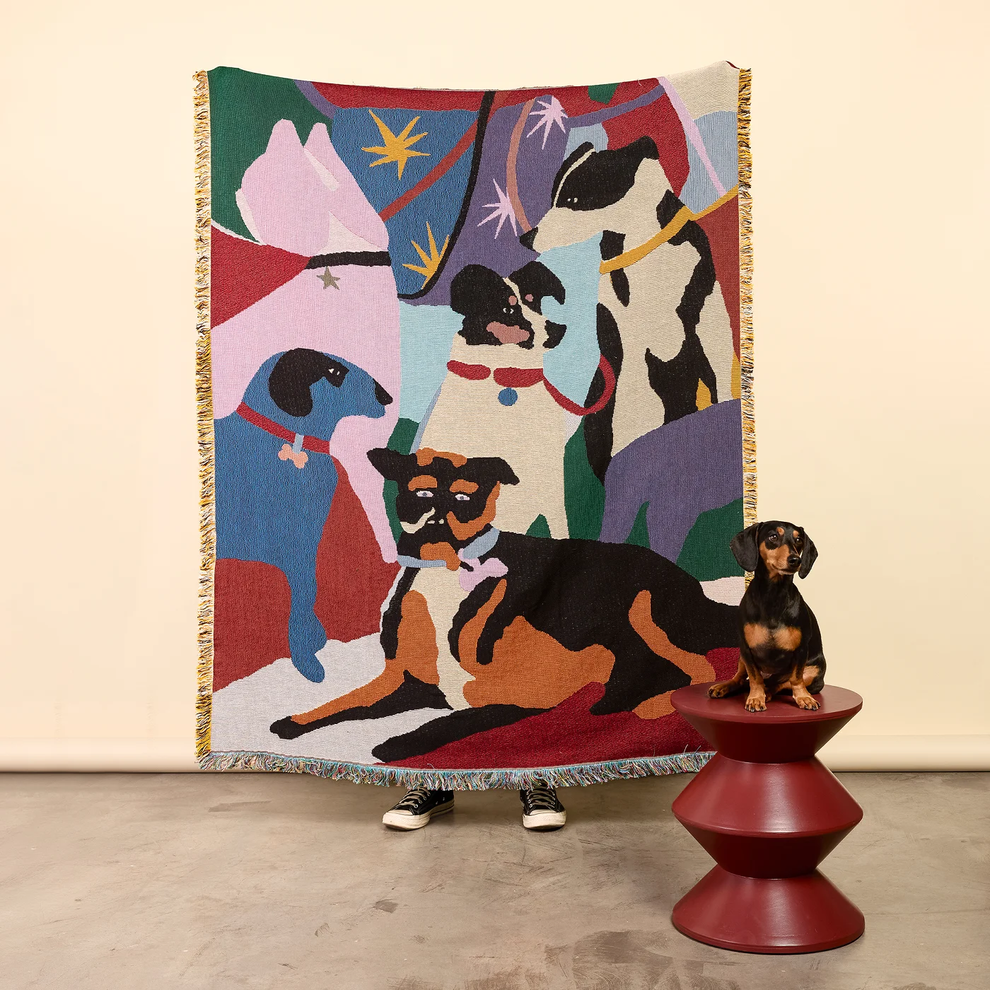 Colorful tapestry with abstract dog figures hanging on wall, person standing behind it, and a small dog statue on a red table.