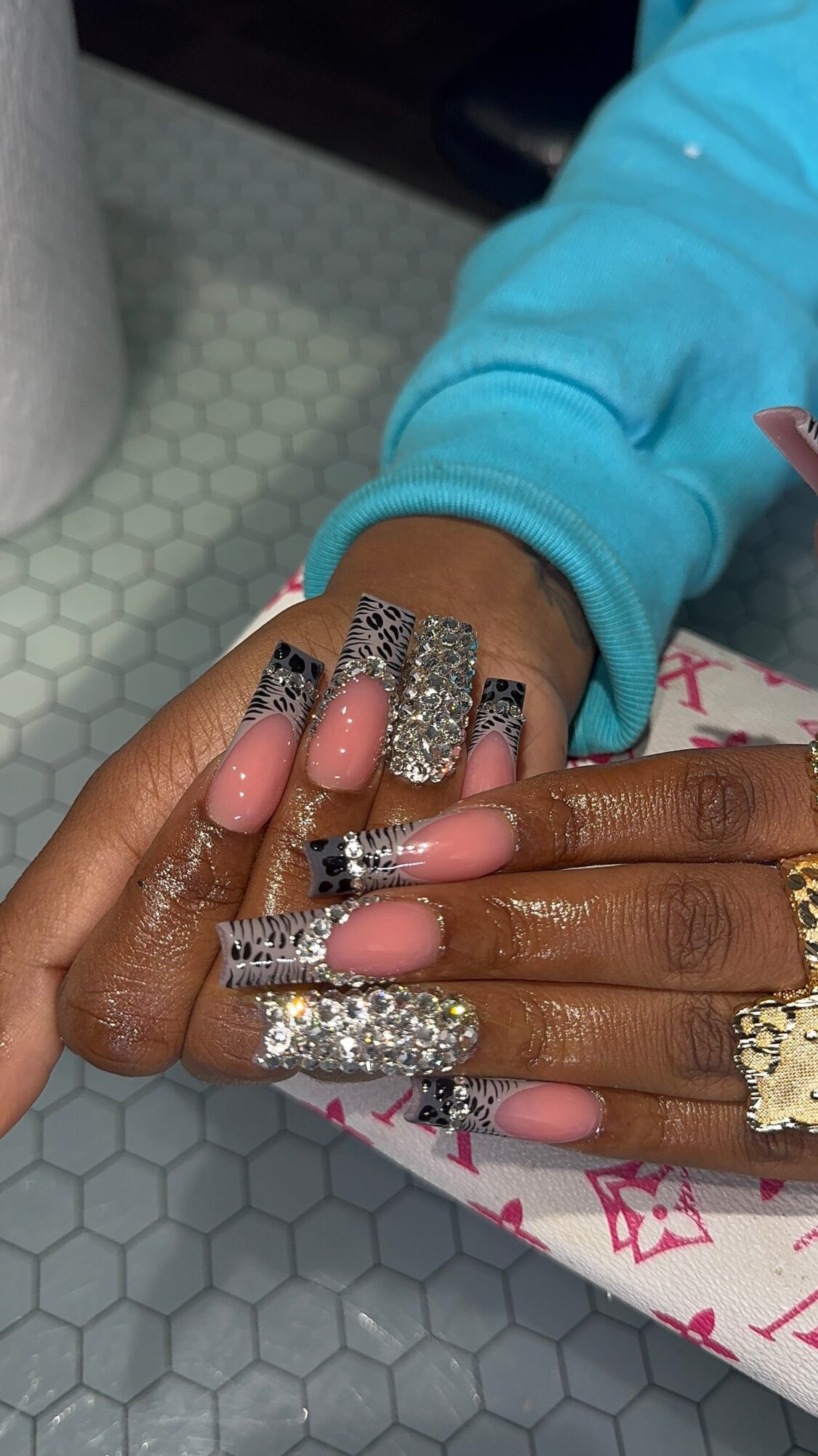 Hands with decorated nails resting on a surface, wearing rings and a blue sleeve, with a patterned background.