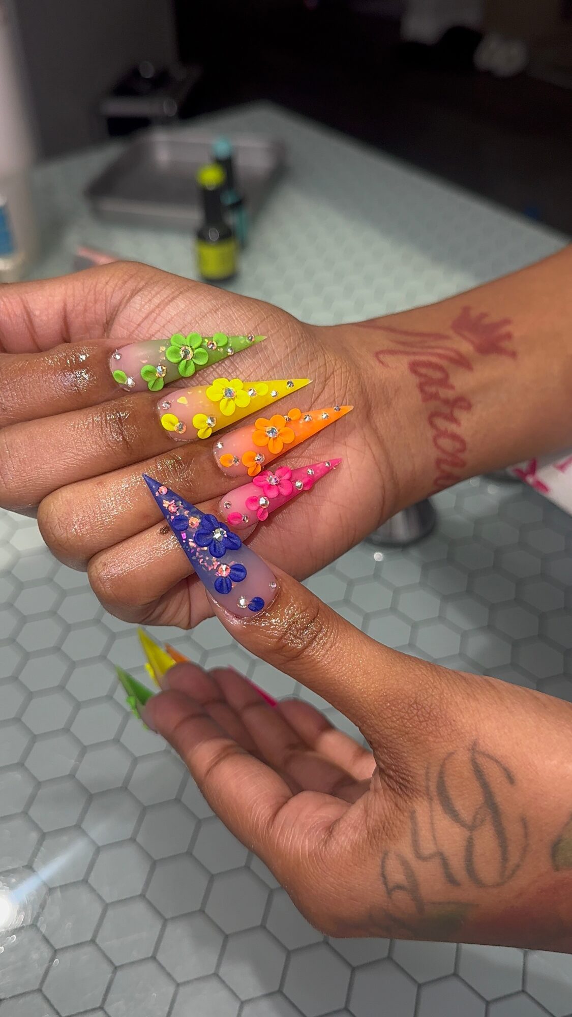 Hands with colorful, decorated long nails and tattoos, displaying nail art with bright colors and rhinestones.