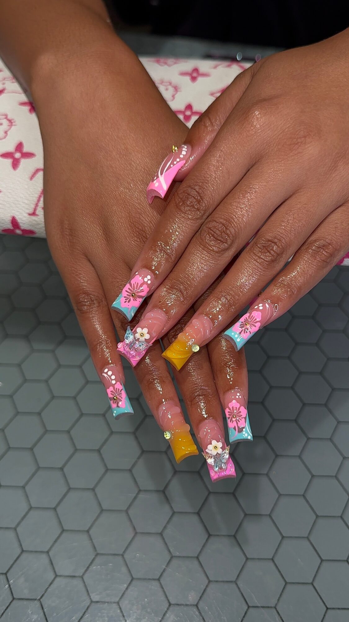 Two hands with long, decorated nails resting on a hexagonal surface, one hand with pink and blue floral nails, the other with yellow tips.