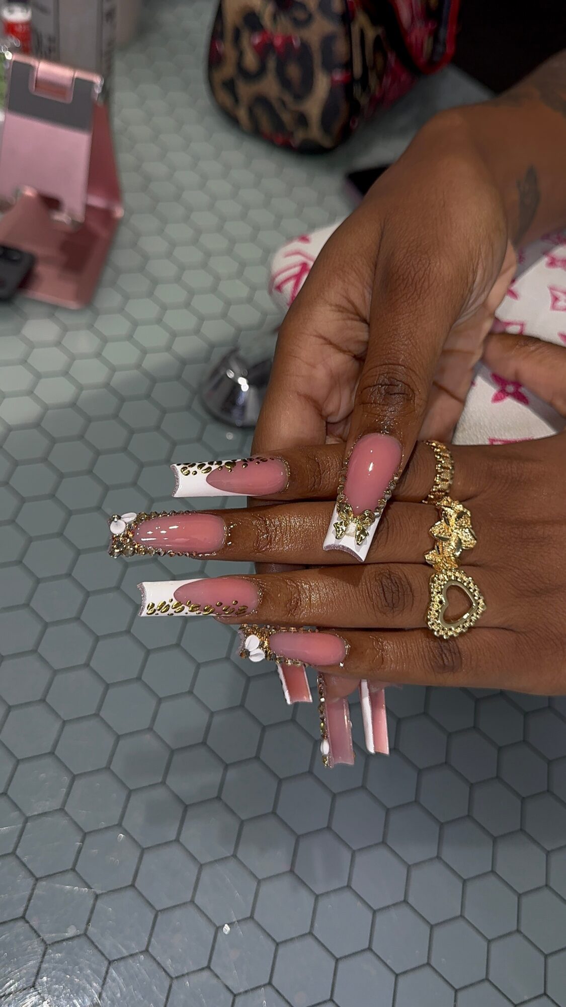 Hands with long, decorated nails and gold jewelry, resting on a hexagonal-tile surface, with a pink box and a leopard-print bag nearby.