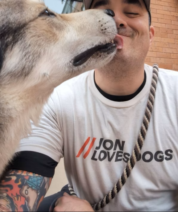 Person smiling with a dog licking their face outdoors, brick wall background, wearing a white T-shirt with text.