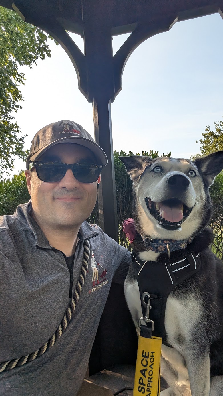 Person wearing sunglasses and a cap taking a selfie with a smiling dog under a shelter, outdoors with trees and blue sky.