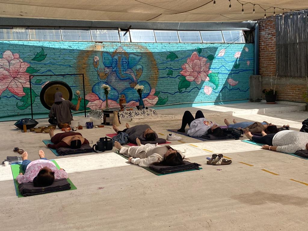People lying on mats in a meditation or yoga session outdoors with a colorful mural of a deity and flowers in the background.