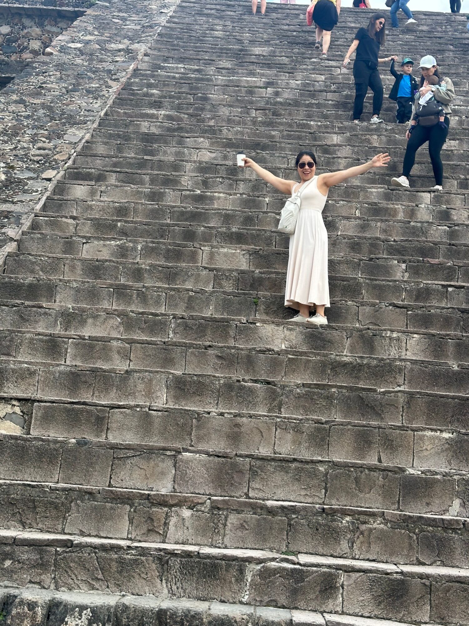 Person in white dress standing on stone steps with arms outstretched, group of people in background, outdoor setting