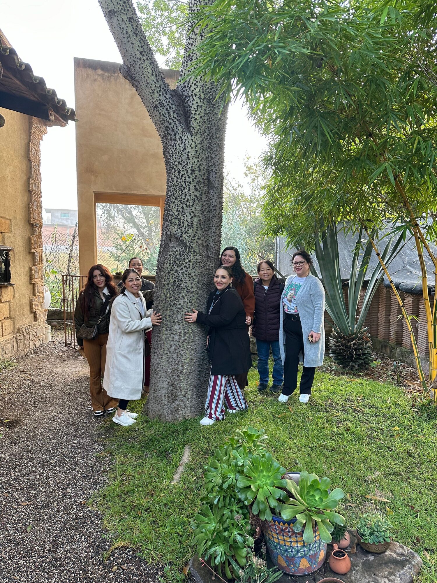 Four people standing outdoors near a tree and a building, with greenery and a gravel path, posing for a photo.