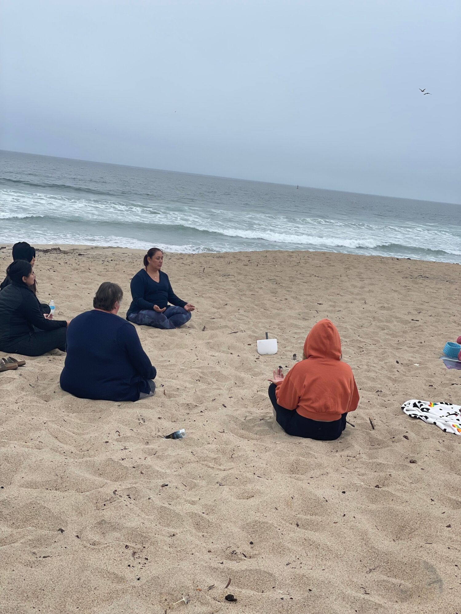 Person sitting on sandy beach facing ocean, wearing a red hoodie, with another person nearby, overcast sky, beach towels visible.