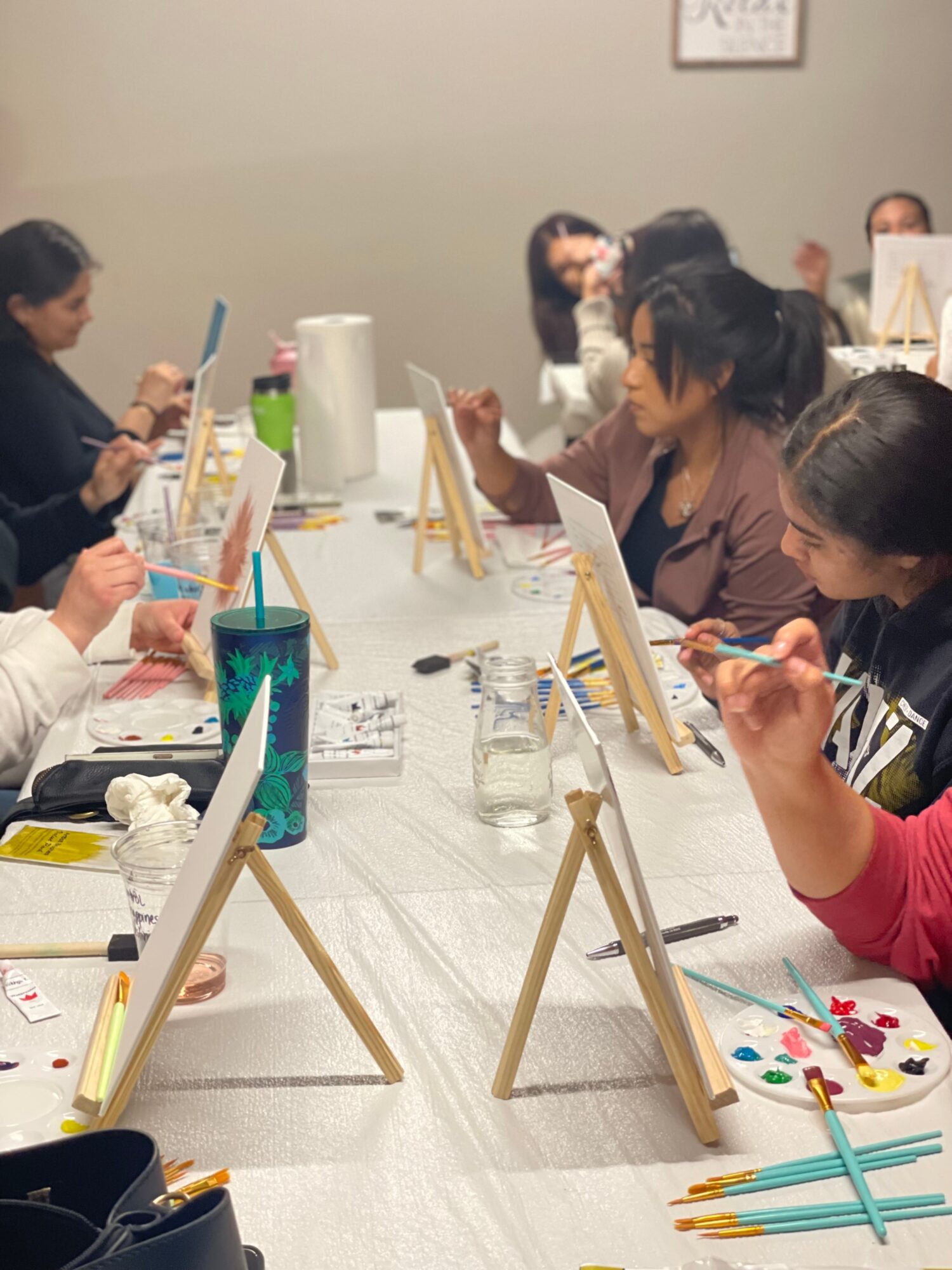 People sitting at a table painting on canvases with brushes, some with palettes, in a room with a gray wall.