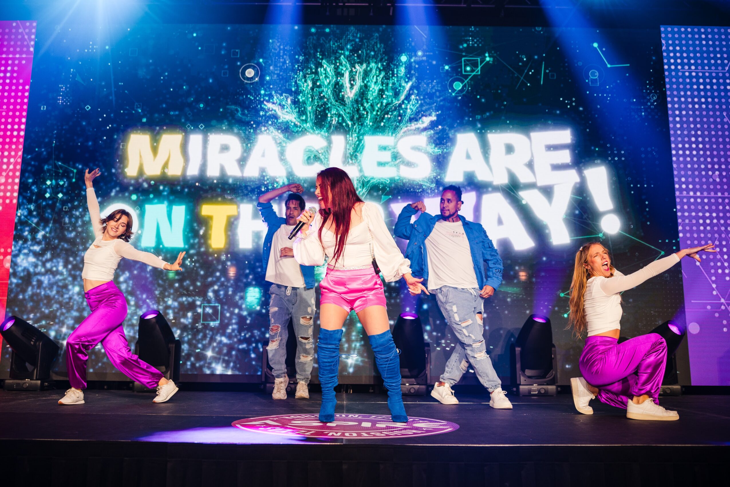 Group of five people dancing on stage with a colorful background and large text reading 'MIRACLES ARE ON THE WAY!'.