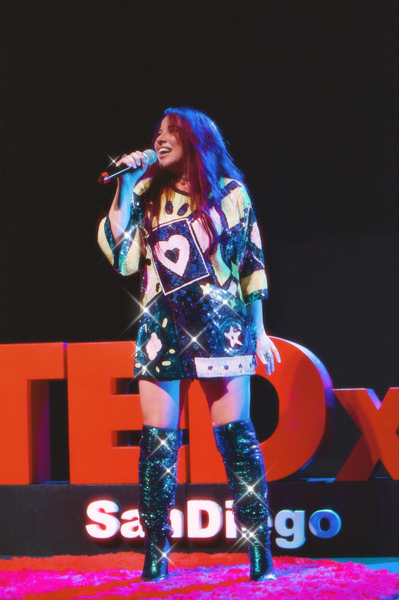 Person singing on stage with large red and white letters spelling 'TEDx' behind them, wearing colorful clothing and knee-high boots.