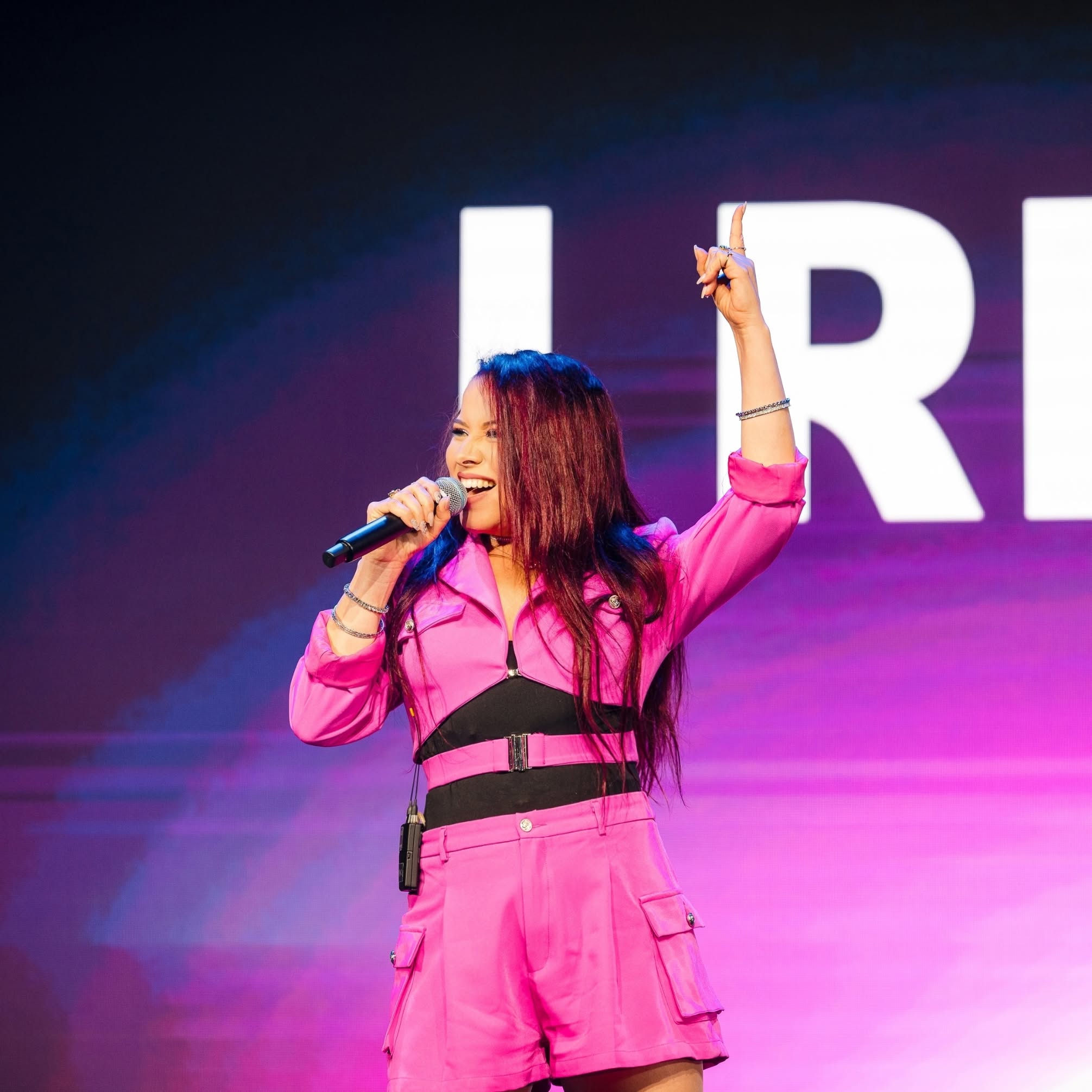 Woman in pink outfit holding microphone, smiling, on stage with large white letters in background.