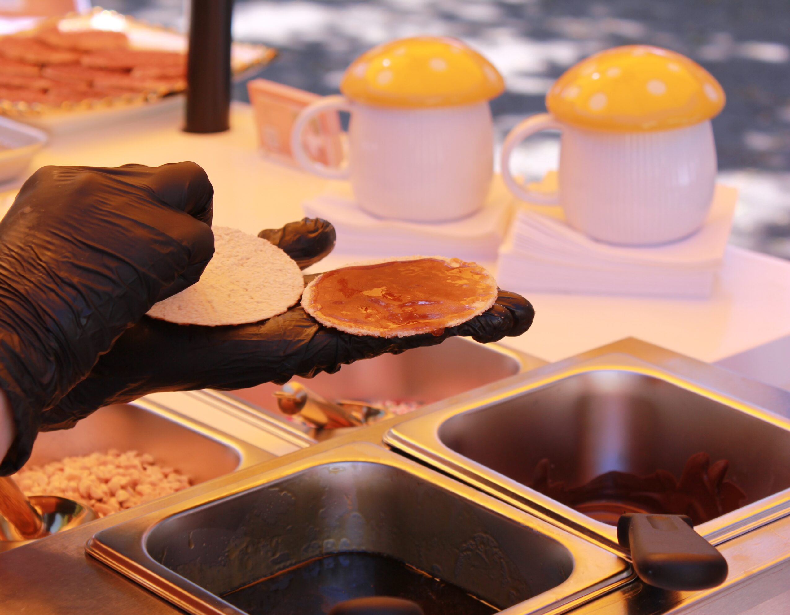 Person wearing black gloves holding a sandwich bun and a slice of tomato, with food containers and mugs in the background.