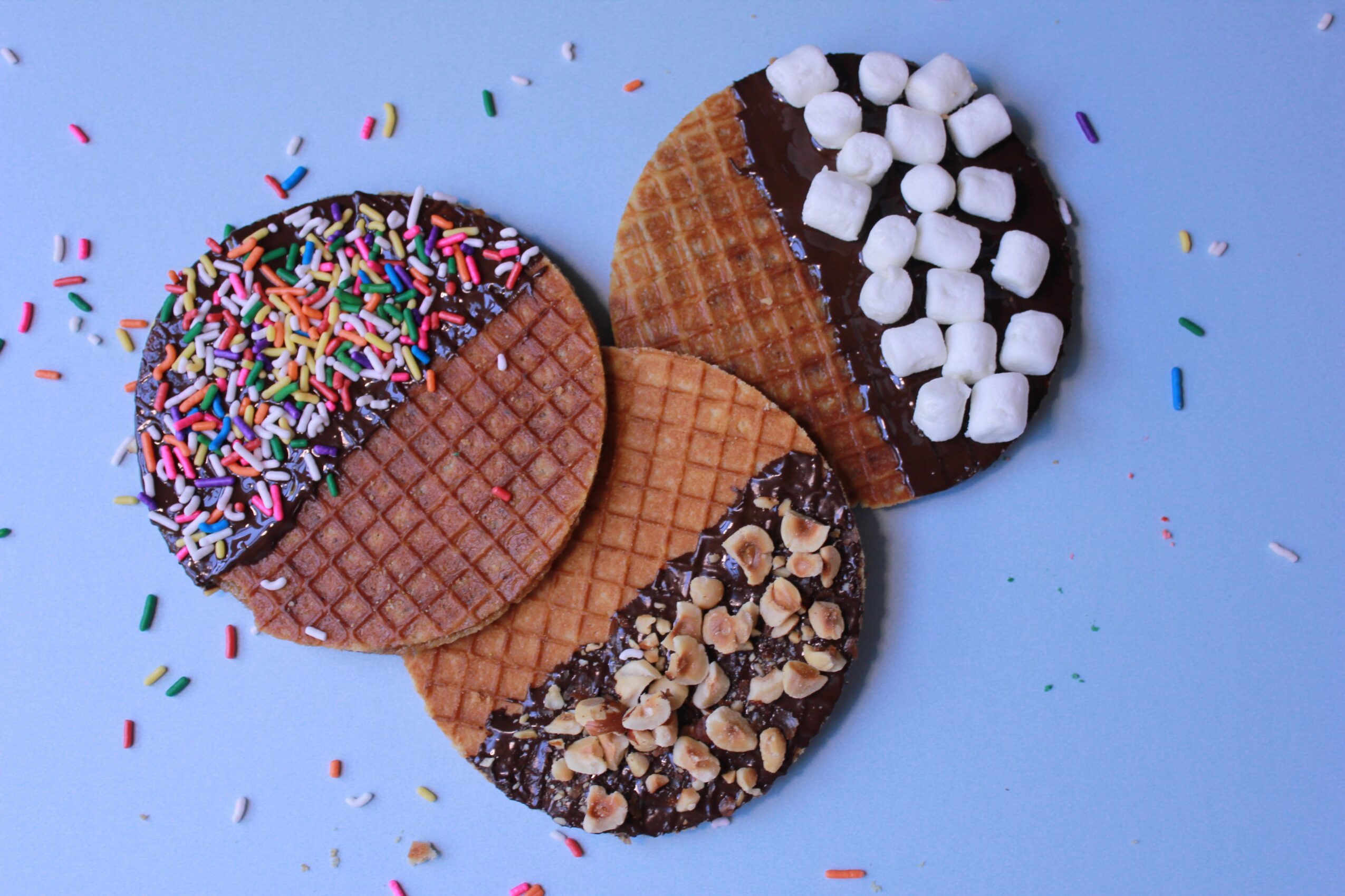 Three waffle cookies with different toppings on a blue surface, surrounded by colorful sprinkles and confetti.
