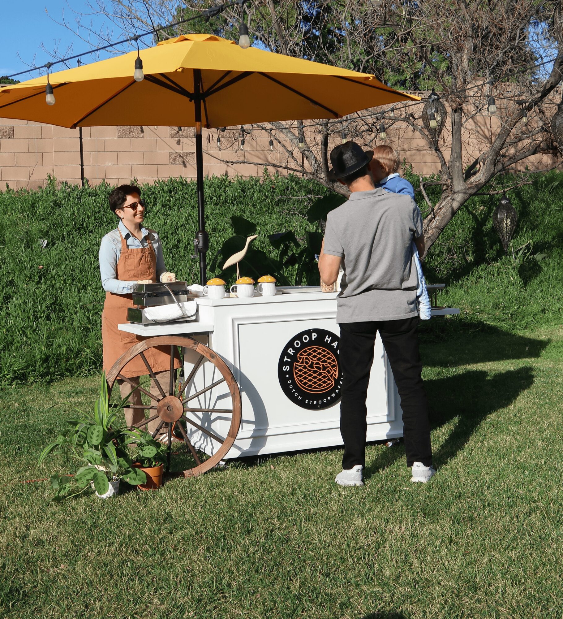 Person serving food at a mobile food cart with a yellow umbrella outdoors, trees and grass in background.