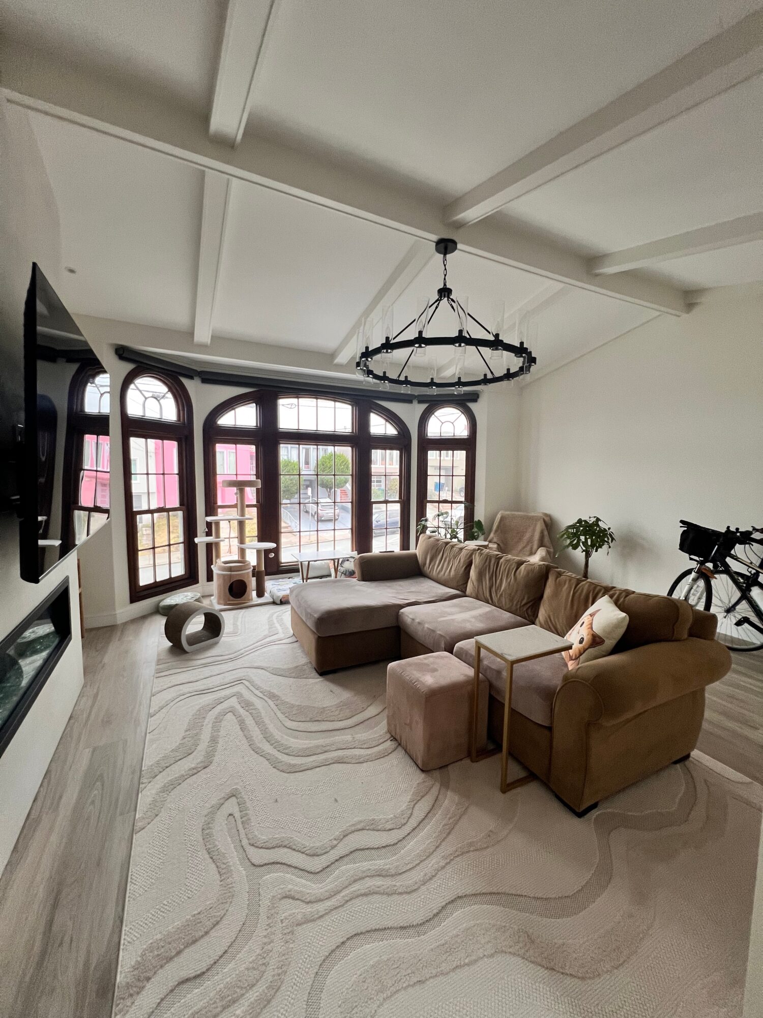 Living room with large arched windows, beige sectional sofa, ottomans, chandelier, and bicycle, with a white rug and potted plant.