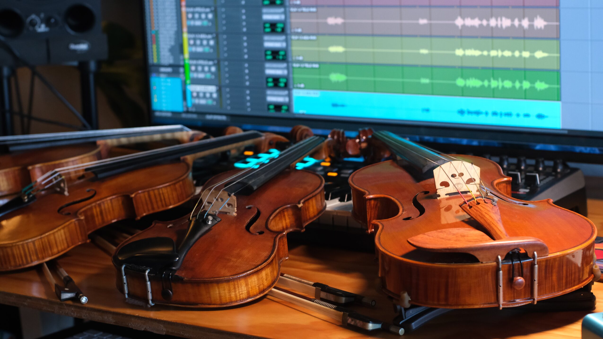 Two violins and a viola on a desk with a computer screen displaying music editing software in the background.
