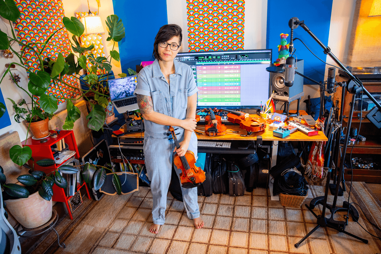 Woman in a recording studio with a computer, microphone, and musical equipment, surrounded by plants and colorful decor.