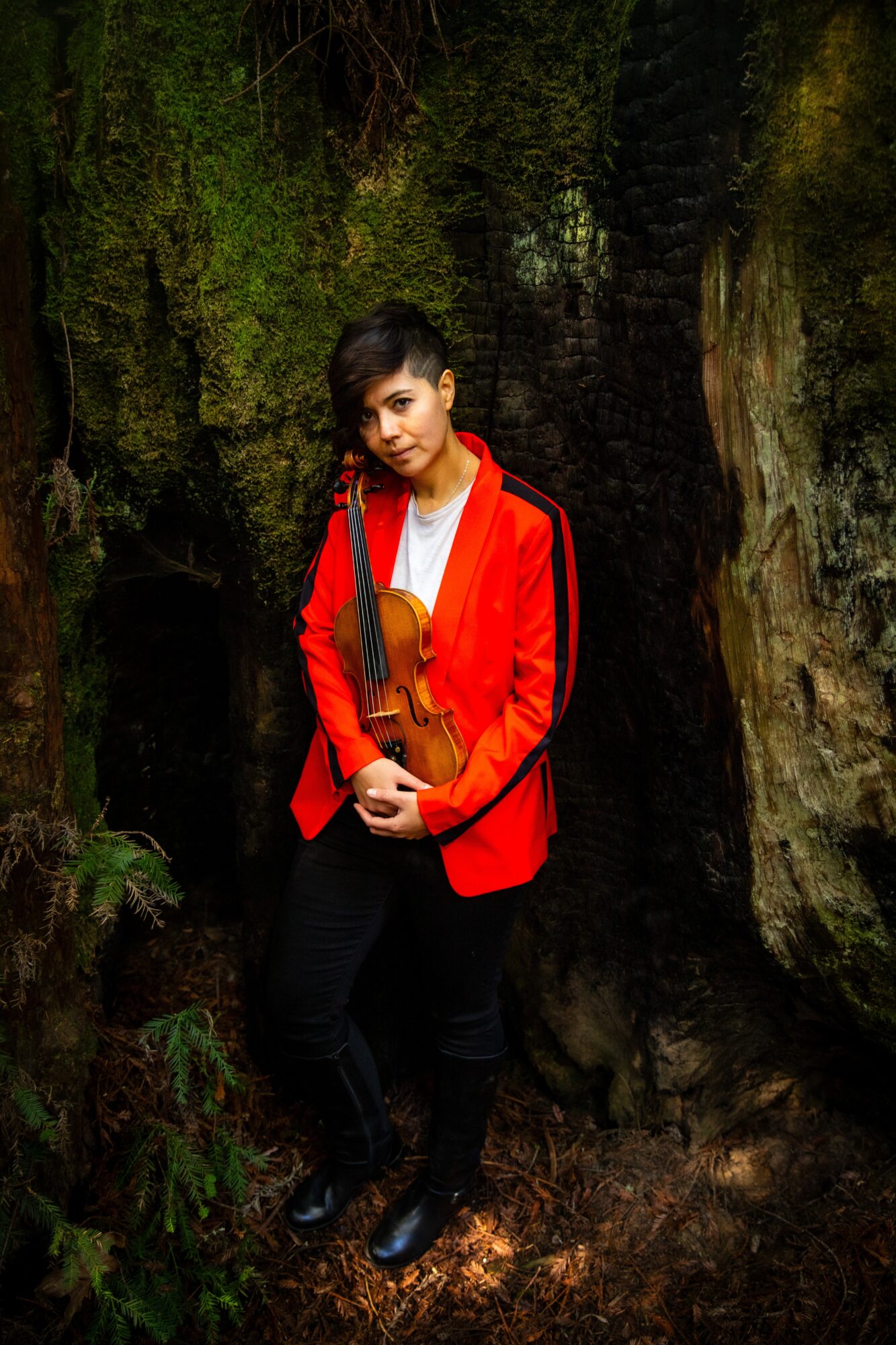 Person holding a violin in a forest, standing between trees, wearing a red jacket and black pants.
