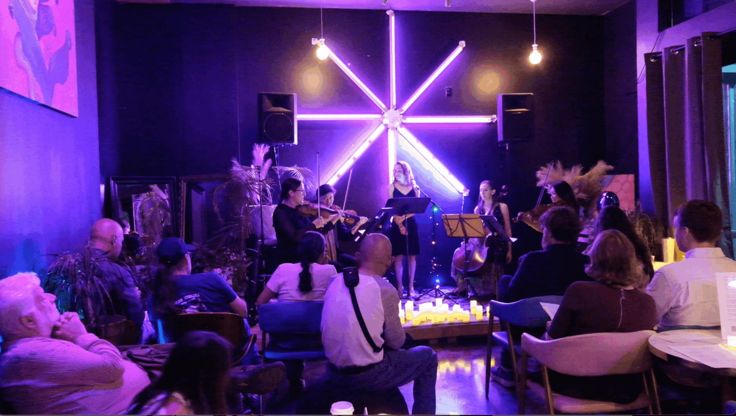 People seated in a dimly lit room watching a musical performance on stage with purple lighting and decorative elements.