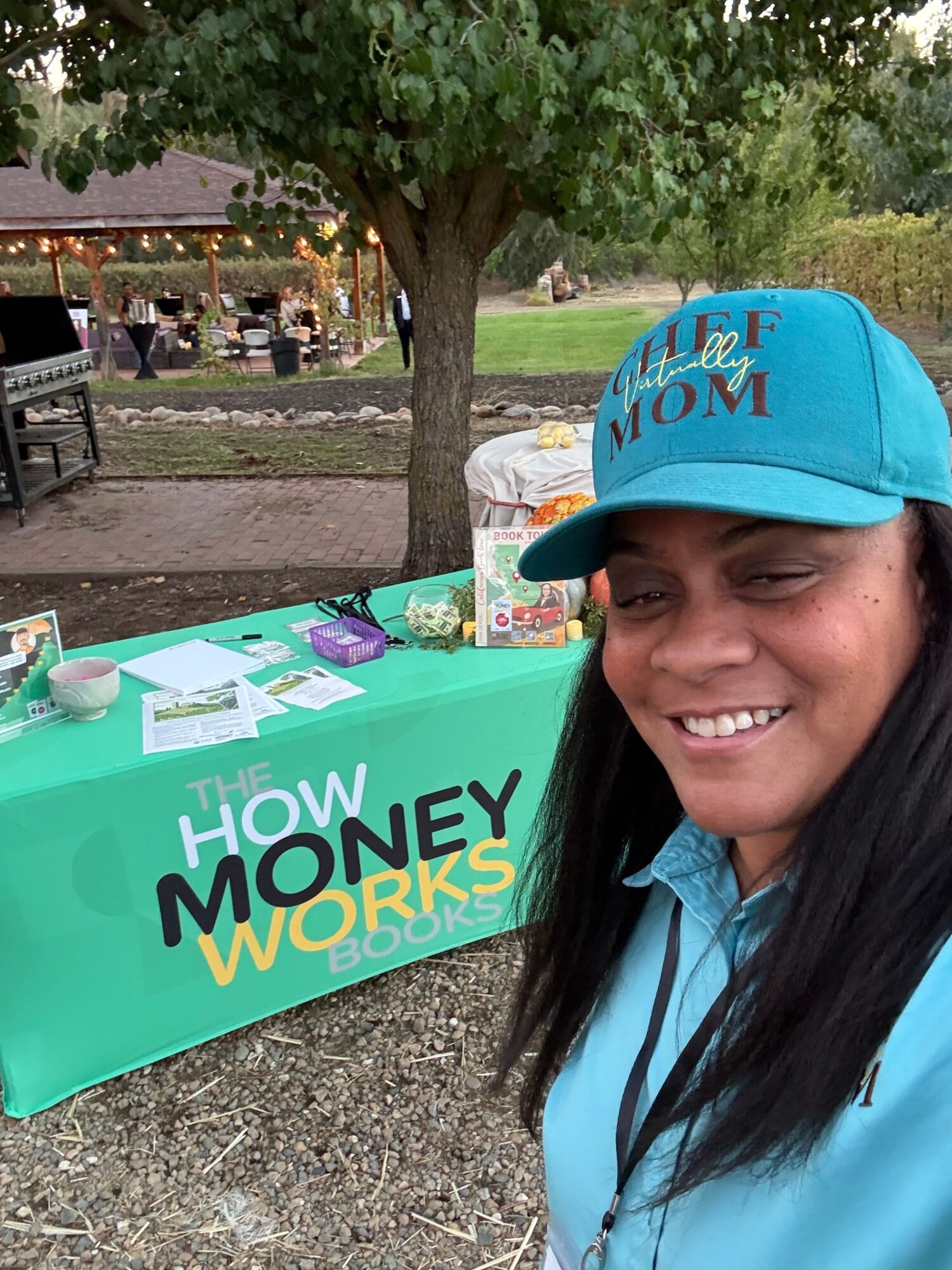 Smiling woman wearing a blue cap and shirt outdoors next to a green table with 'How Money Works' sign.