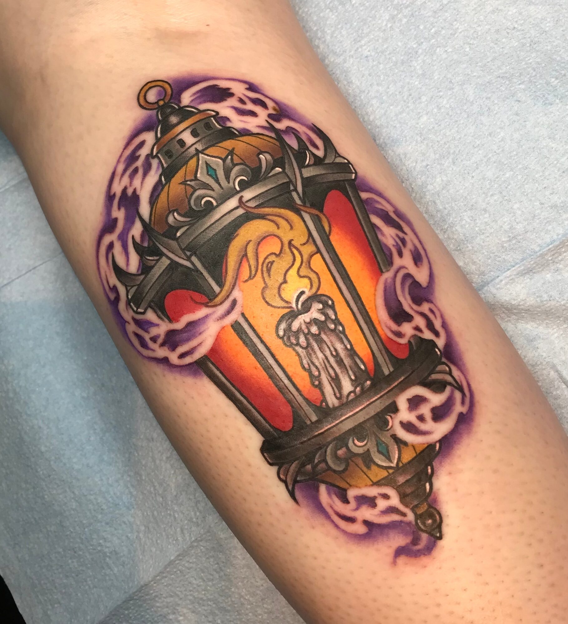 Tattoo of a lantern with flames and purple smoke, detailed with metallic and fiery elements, on skin.