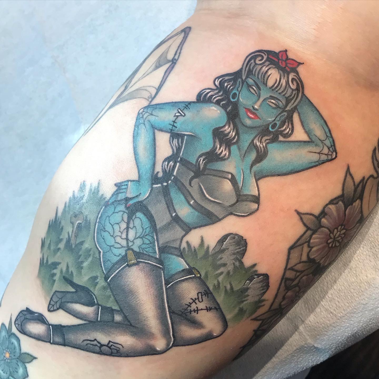 Tattoo of a woman with blue skin, long dark hair, and devil horns, kneeling outdoors with plants in the background.