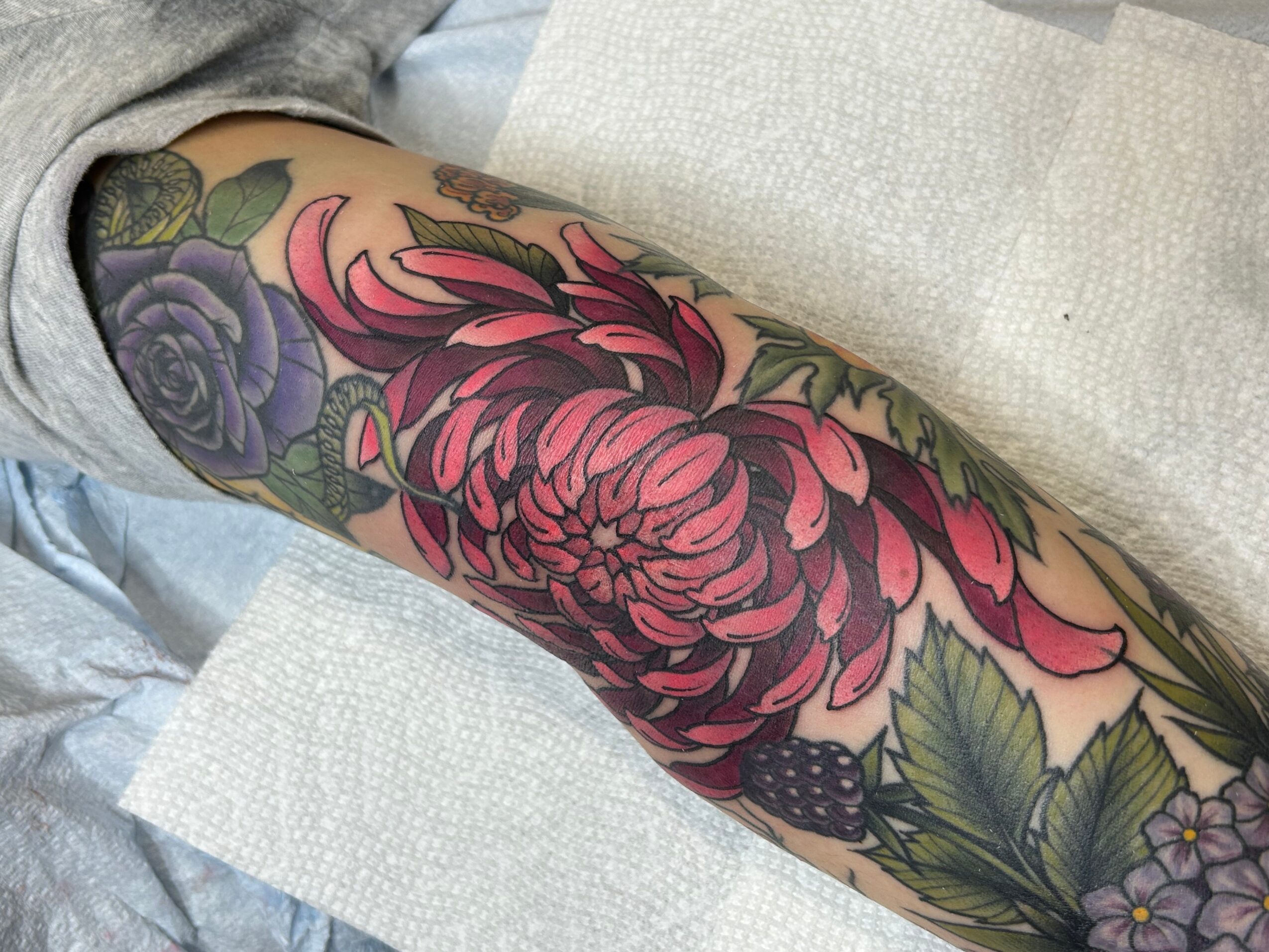 Tattooed arm with a large pink flower, purple roses, and green leaves, resting on a white surface.