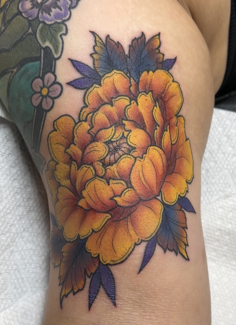Colorful peony flower tattoo with orange and yellow petals and dark blue leaves on skin.
