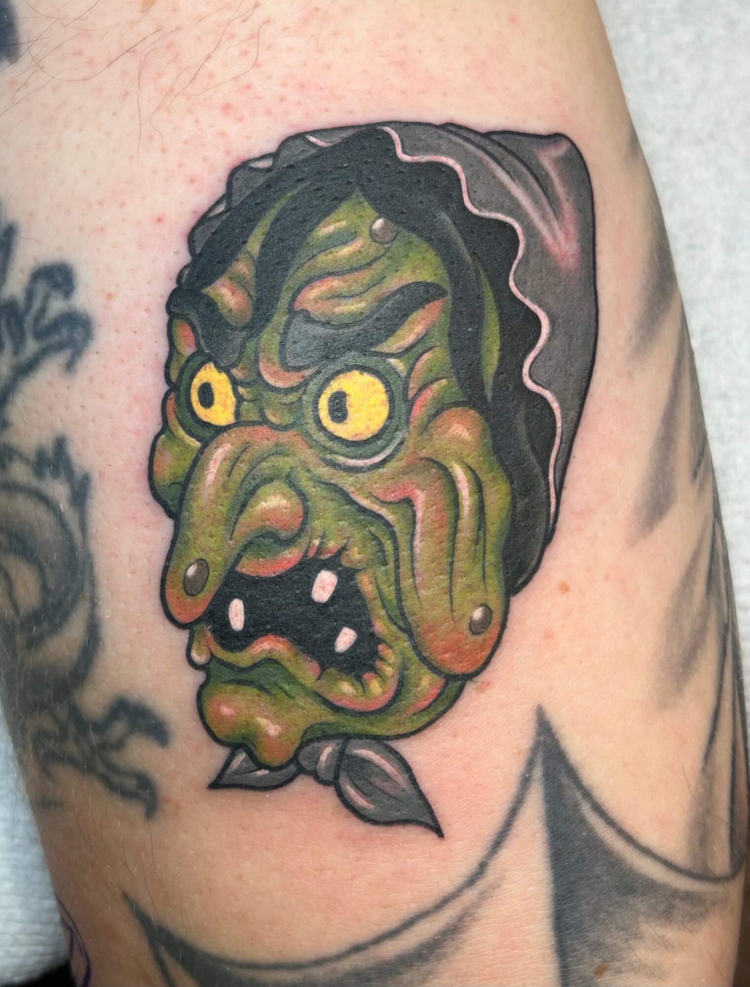 Colorful witch face tattoo with green skin, yellow eyes, and a black hat, on skin with other tattoos visible.