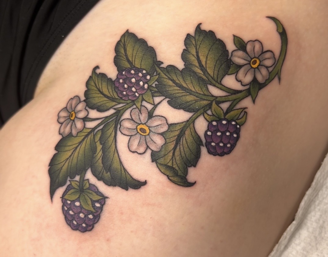 Tattoo of blackberry plant with green leaves, white flowers, and blackberries on skin.