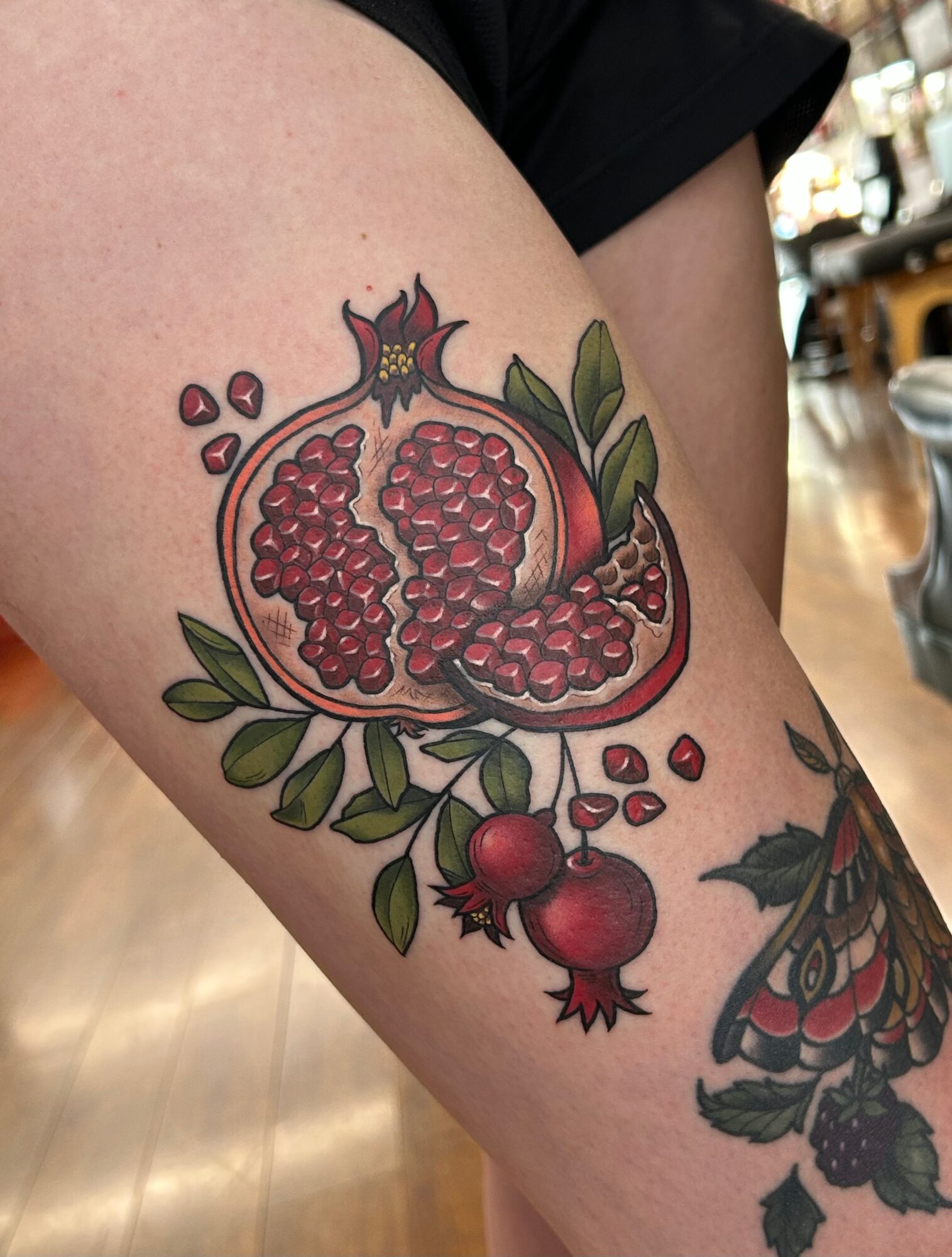 Tattoo of a pomegranate with leaves and a partially opened fruit showing seeds, on a person's arm.