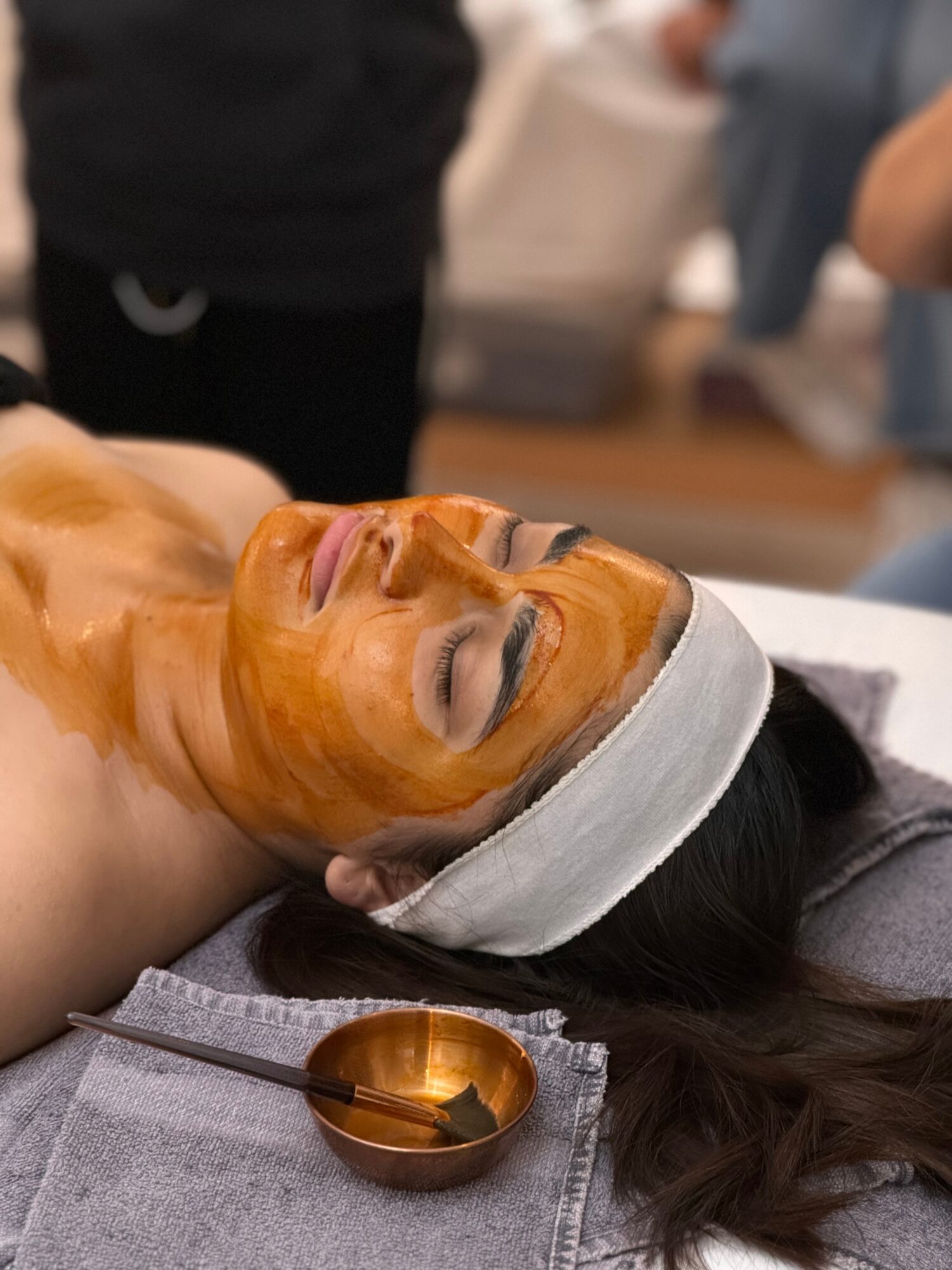 Person receiving facial treatment with a mask, lying down with eyes closed, head on a towel, in a spa setting.