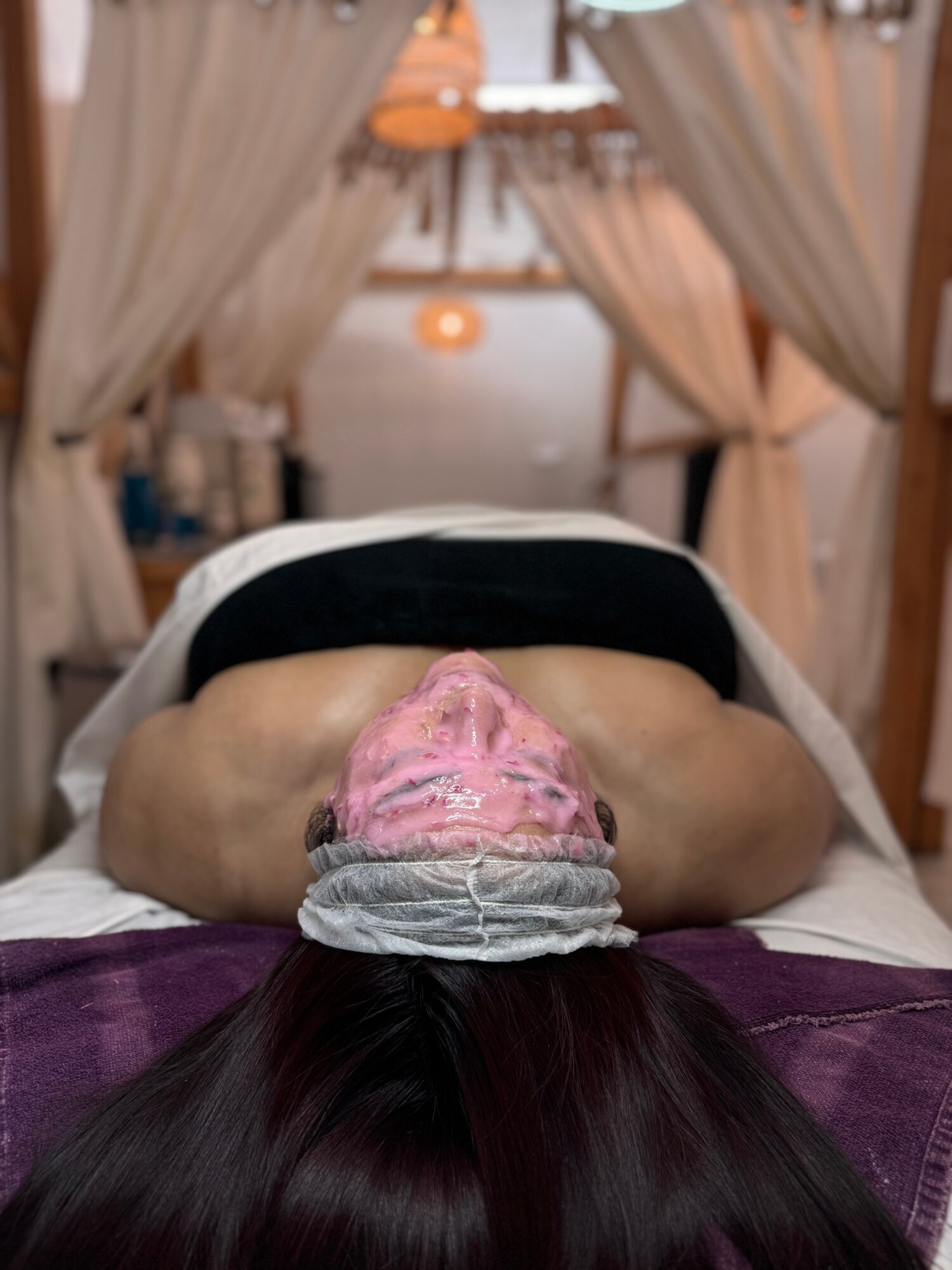 Person lying face down on a massage table with a towel on their head, in a room with curtains and warm lighting.