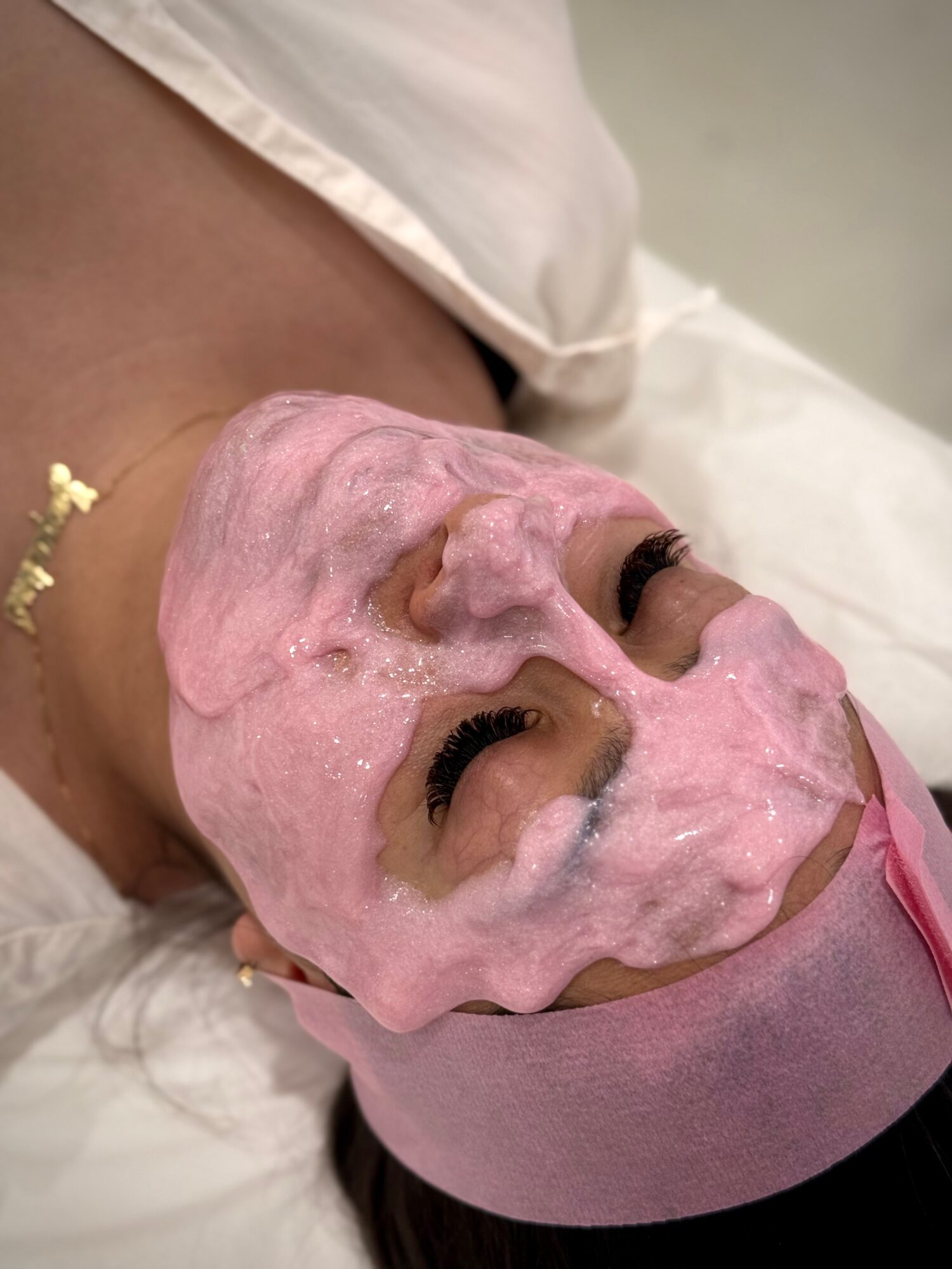 Person lying down with a pink facial mask, eyes closed, wearing a headband, in a spa or skincare setting.
