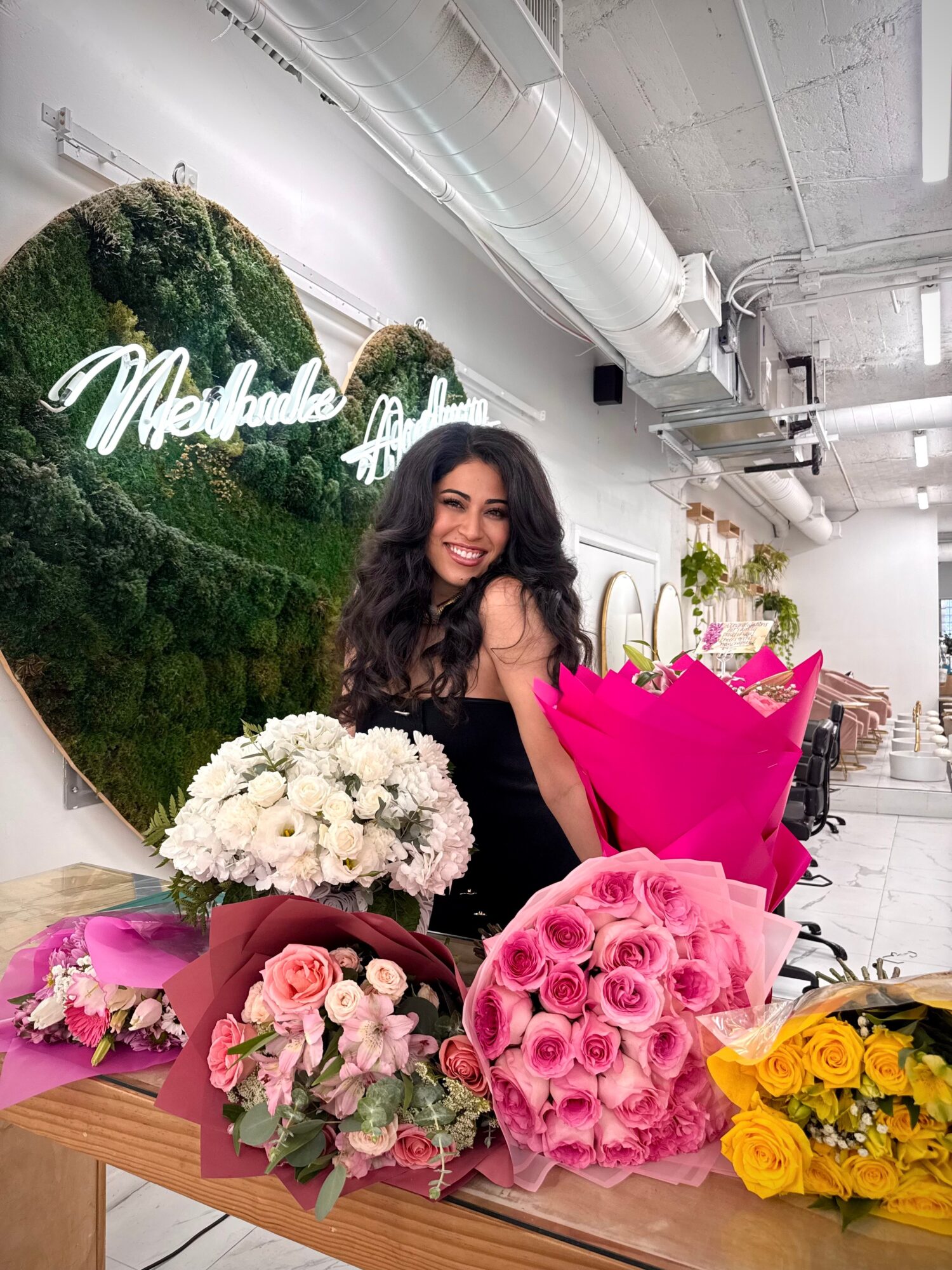 Smiling woman with long dark hair surrounded by colorful flower bouquets in a room with a green moss wall and neon sign.