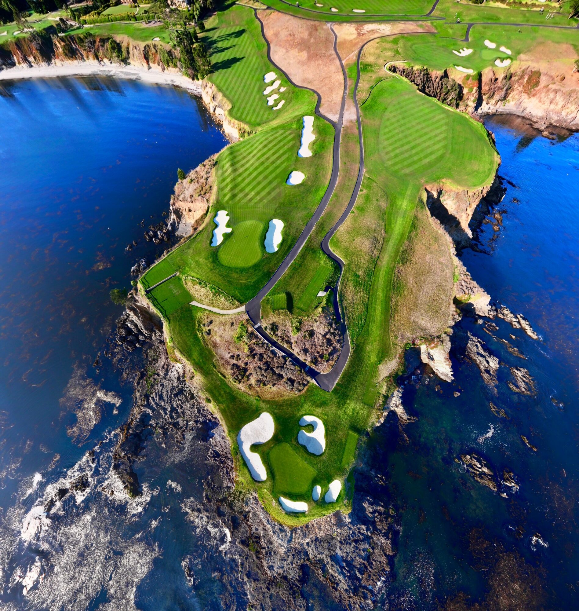 Aerial view of a golf course on a cliffside with water on three sides, featuring green fairways, sand traps, and pathways.