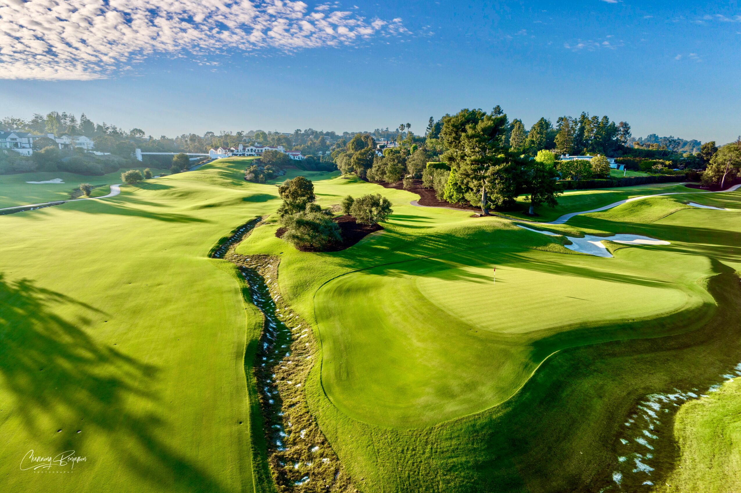 Golf course with green fairways, sand traps, trees, and a blue sky with clouds.