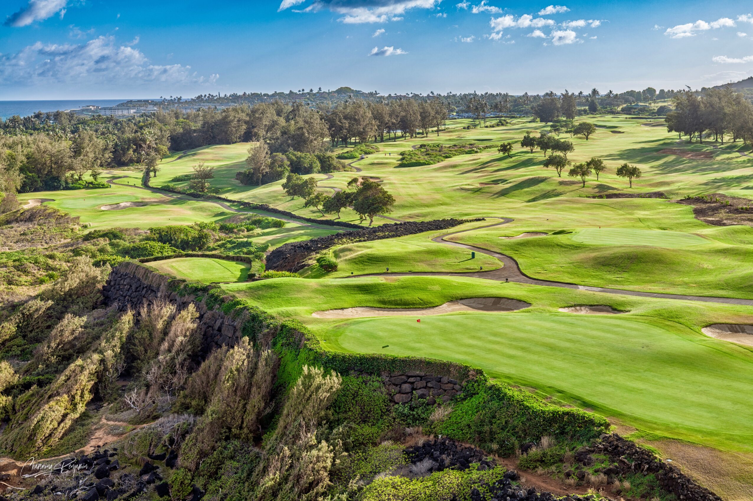 Golf course with green fairways, sand traps, and trees under a blue sky with clouds.