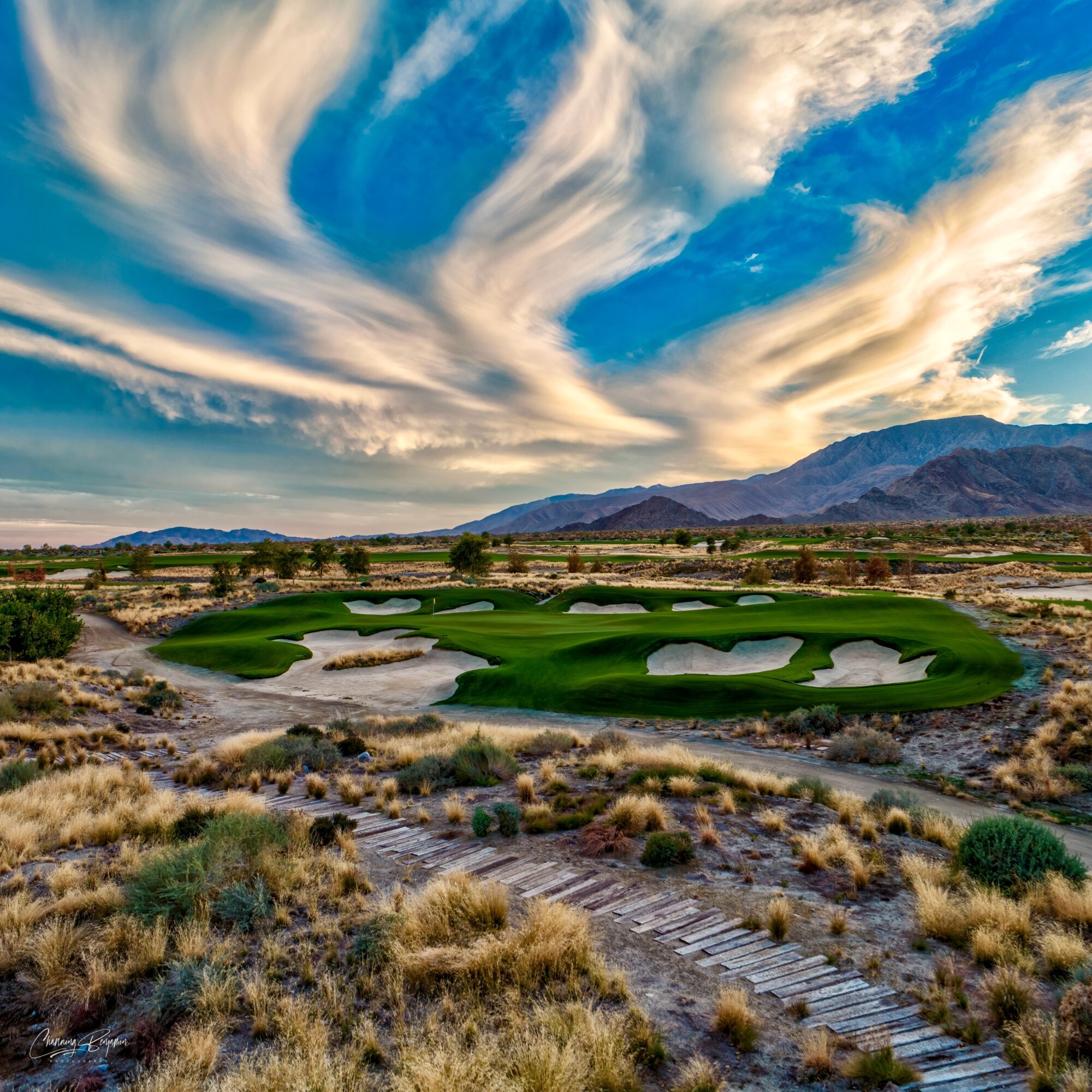 Desert landscape with a golf course, mountains in the background, and dramatic clouds in the sky.