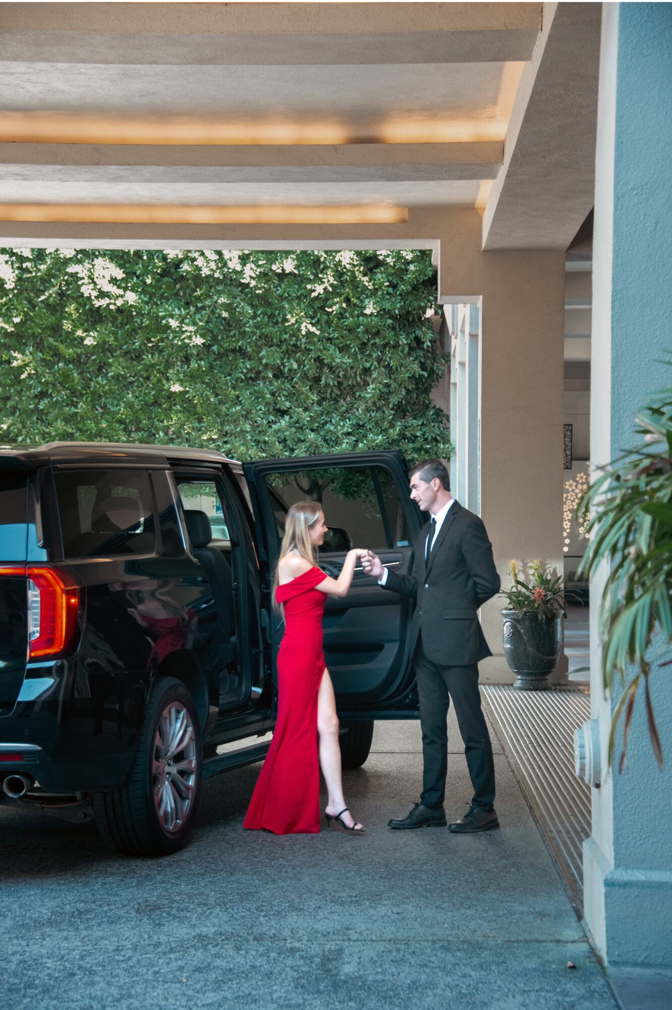 A woman in a red dress and a man in a suit are exchanging a small item outside a building with black vehicles nearby.