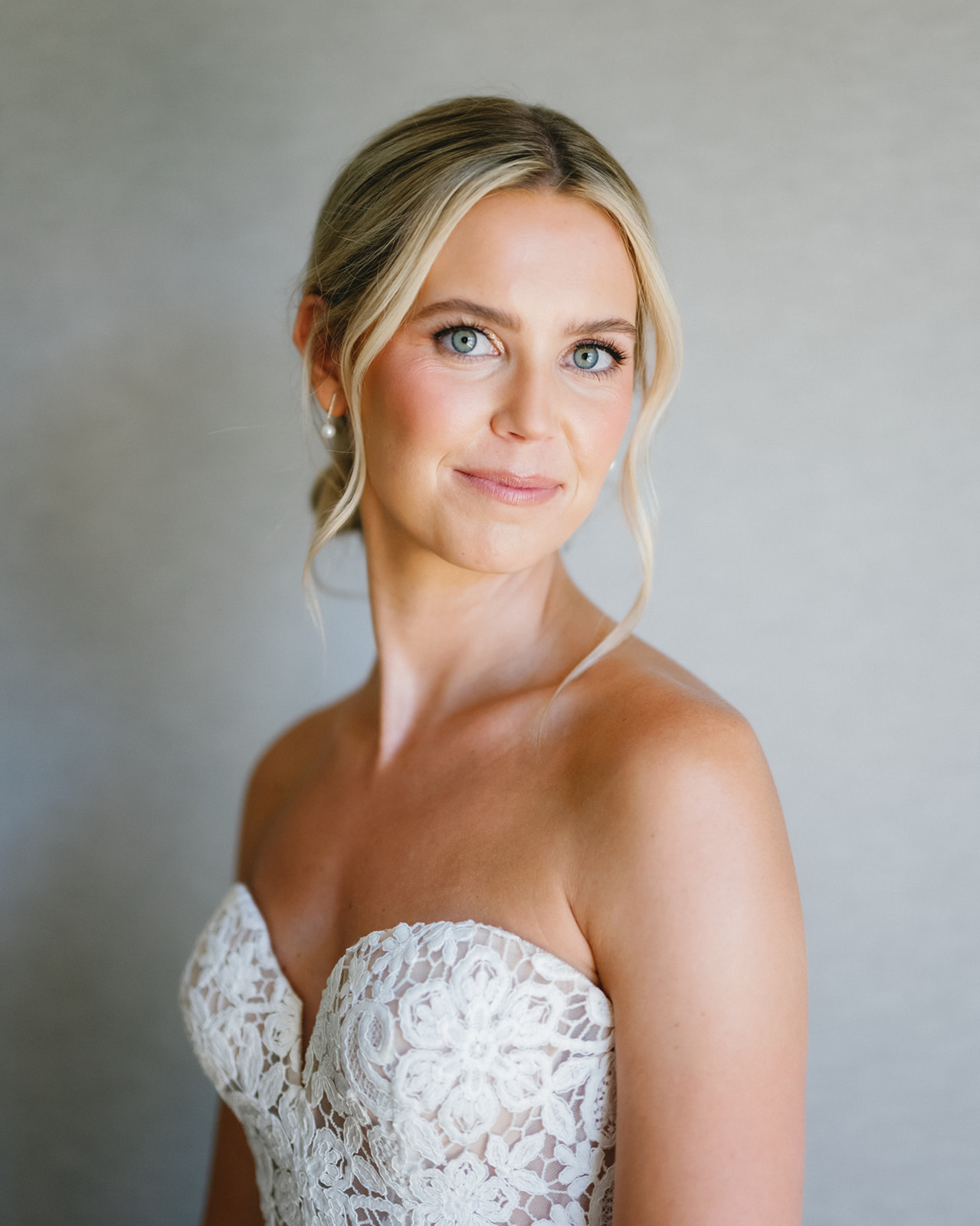Portrait of a woman with blonde hair in a strapless lace dress, smiling softly, against a plain background.