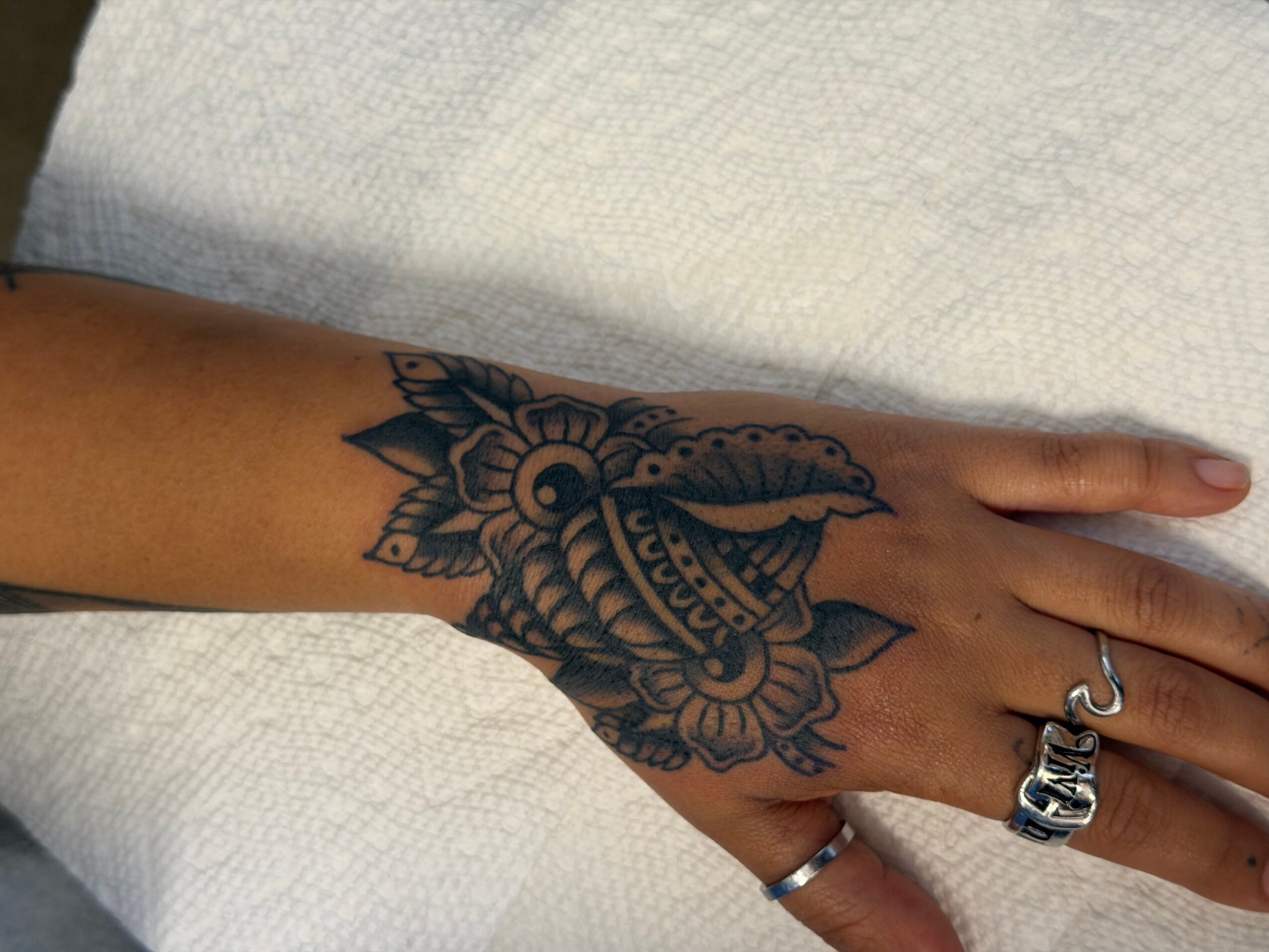 Hand with a detailed black and gray floral tattoo on the back, wearing rings, resting on a light-colored surface.