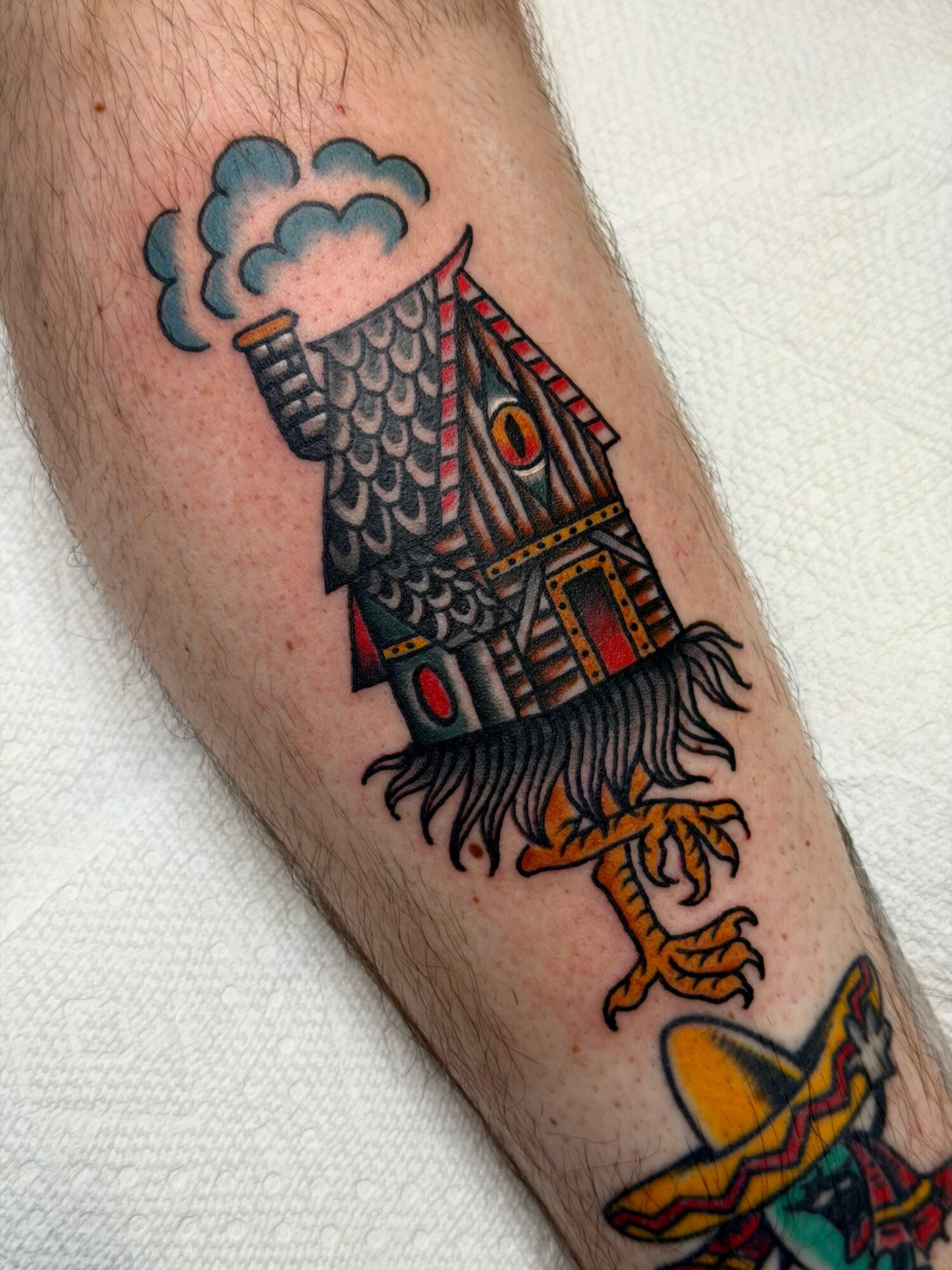 Tattoo of a house with a smoking chimney, a tiger, and a colorful object, all in a cartoon style.