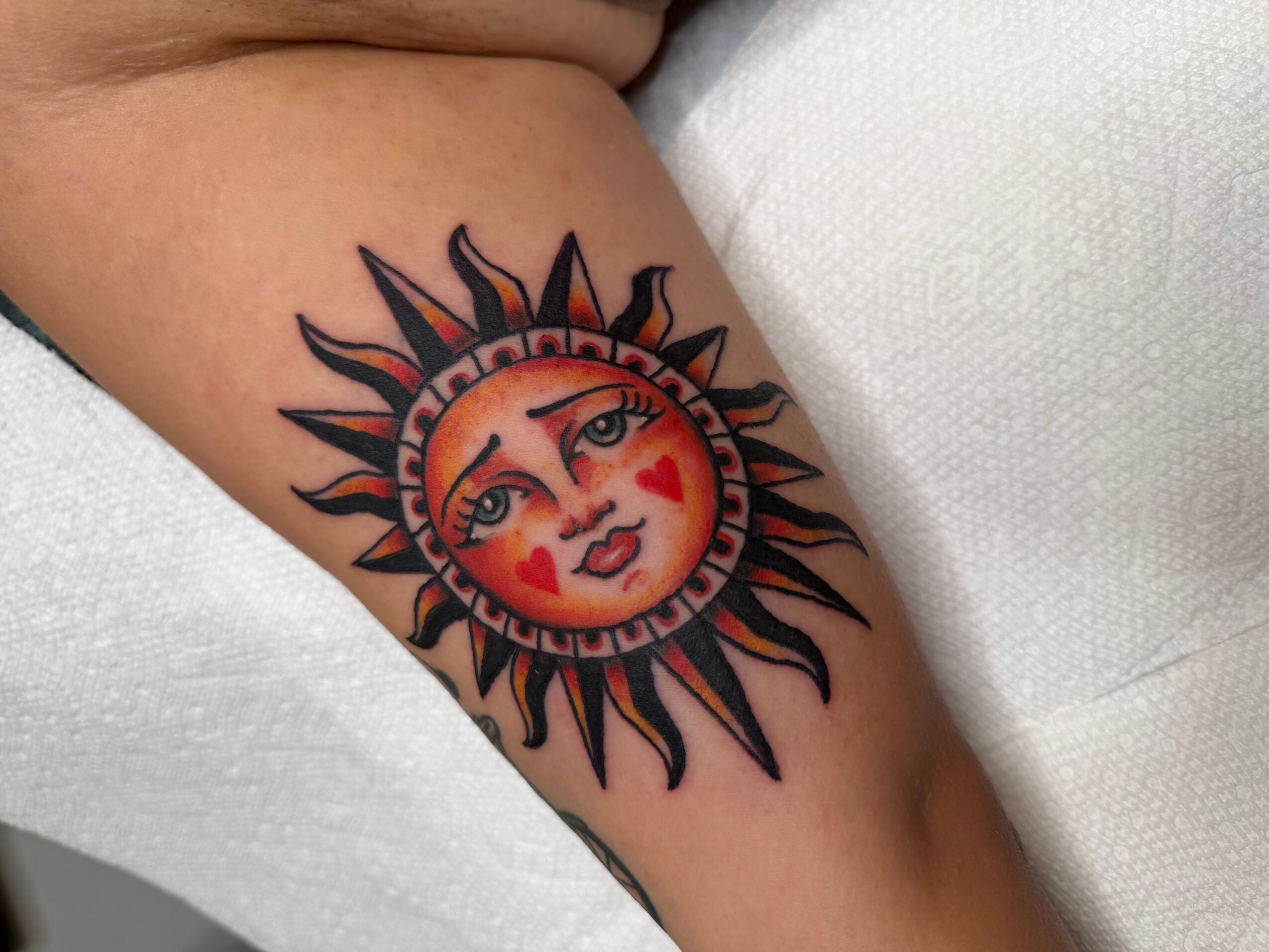 Tattoo of a sun with a face, featuring a winking eye, red lips, and red hearts on cheeks, surrounded by black and orange rays.