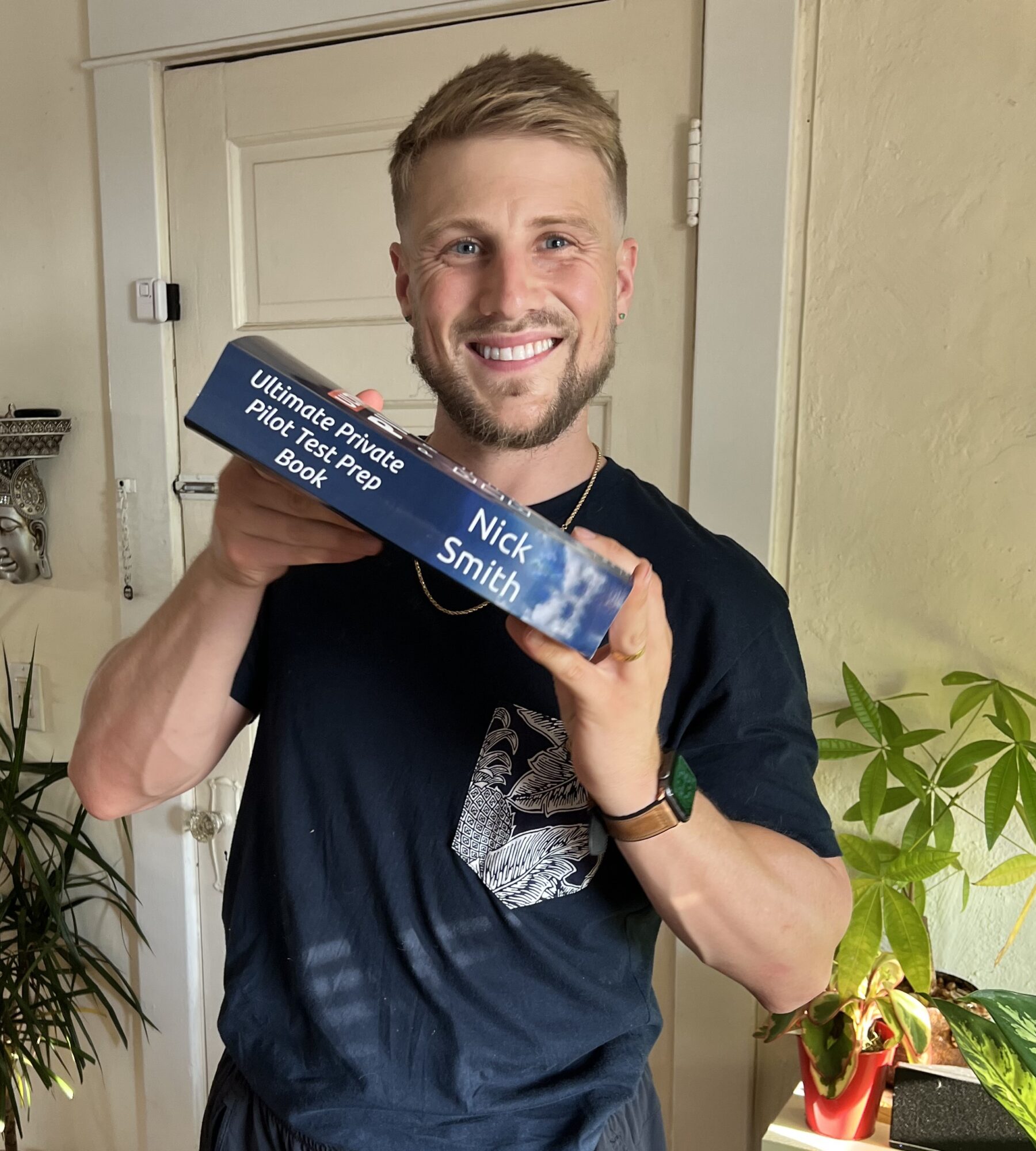 Smiling man holding a blue box with text, standing indoors near a door and plants.